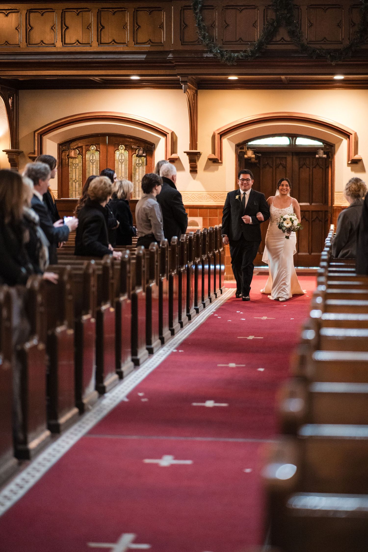 A couple walks down a grand church aisle with wooden pews and a red carpet runner.