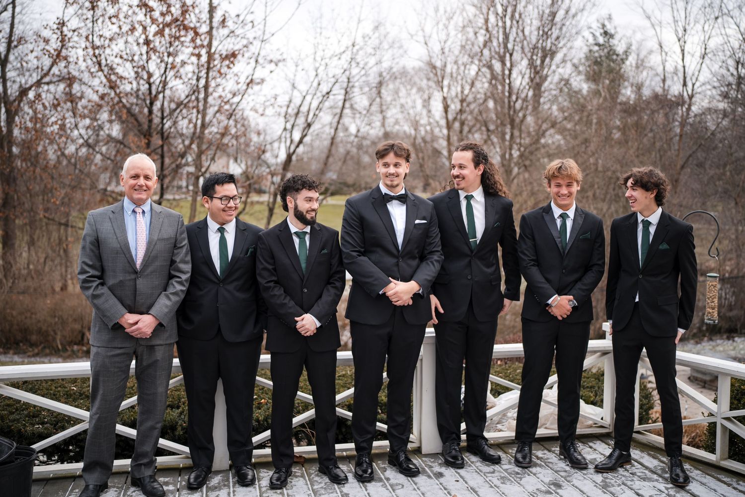 Wedding party in formal black suits standing together on a wooden deck during winter.