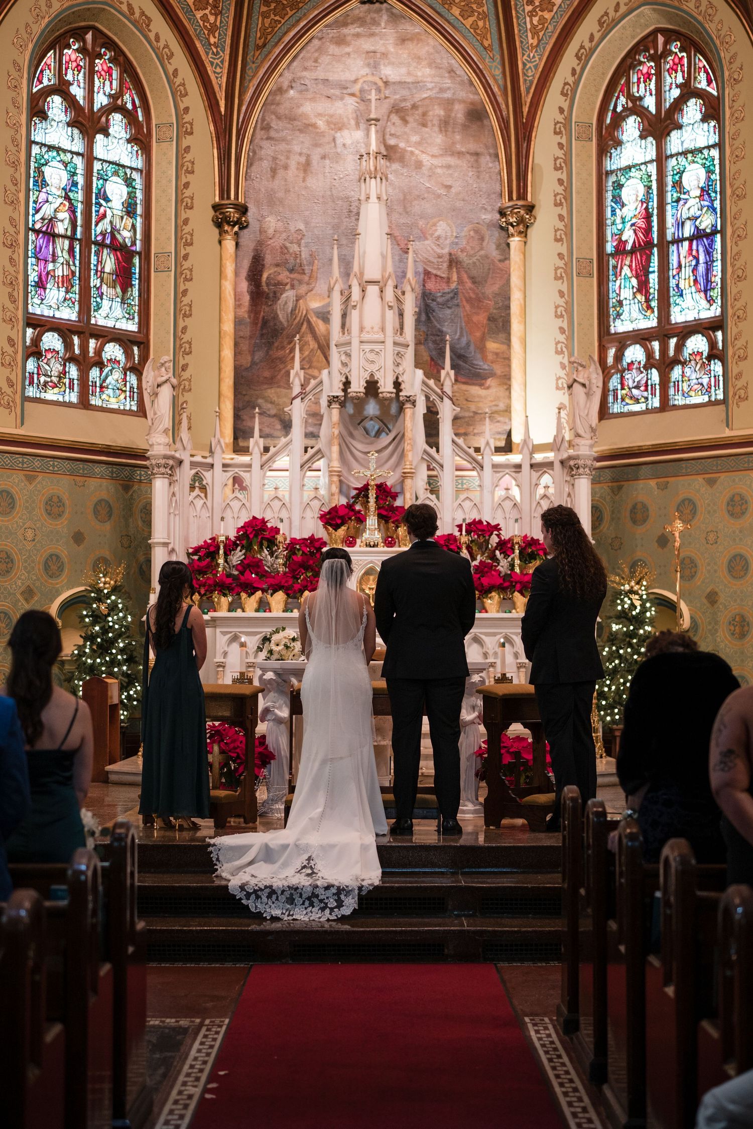 A wedding ceremony takes place before an ornate altar decorated with red poinsettias and stained glass windows.