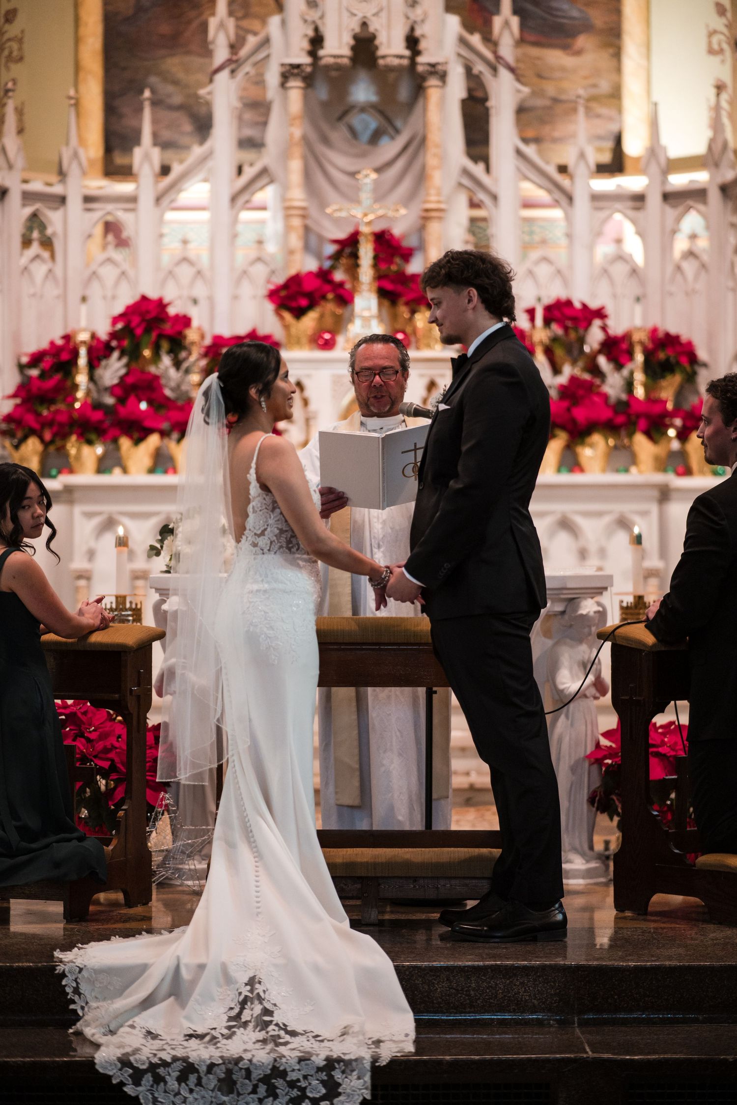 A couple holds hands at the altar during their wedding ceremony surrounded by red poinsettias.
