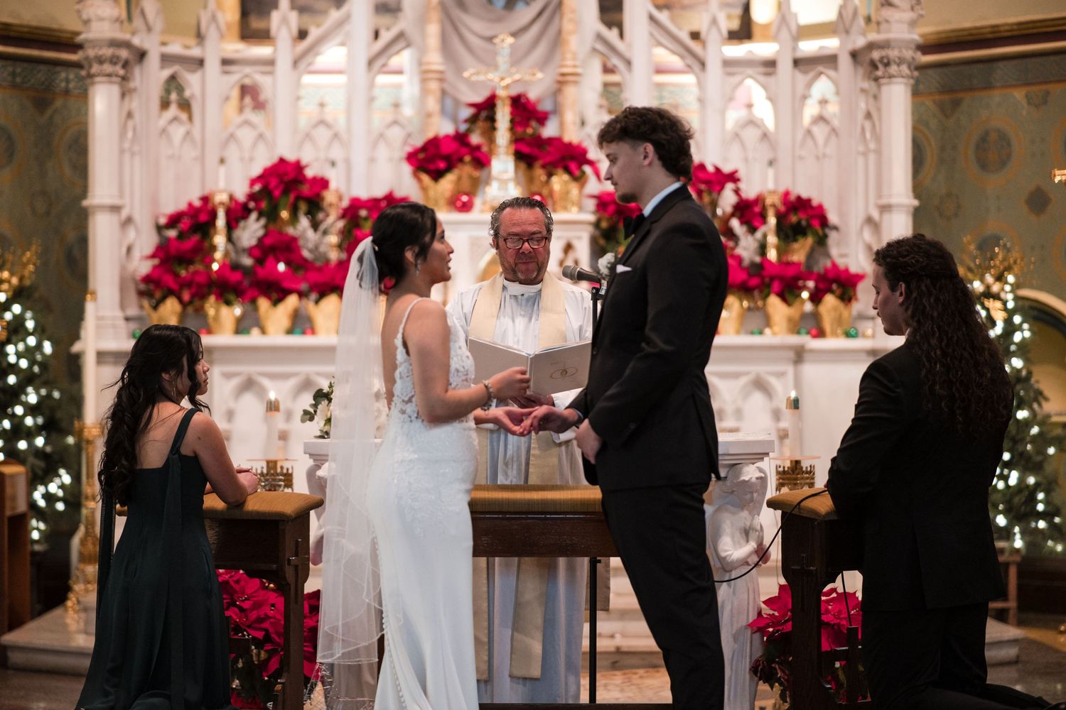 A wedding ceremony in progress at an ornate altar decorated with vibrant red poinsettias and Christmas lights.