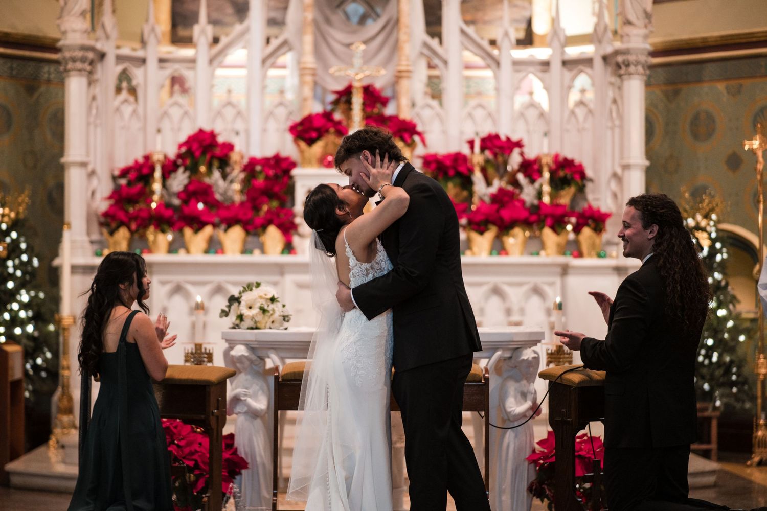 Newlyweds share their first kiss as a married couple in front of an altar adorned with red flowers.