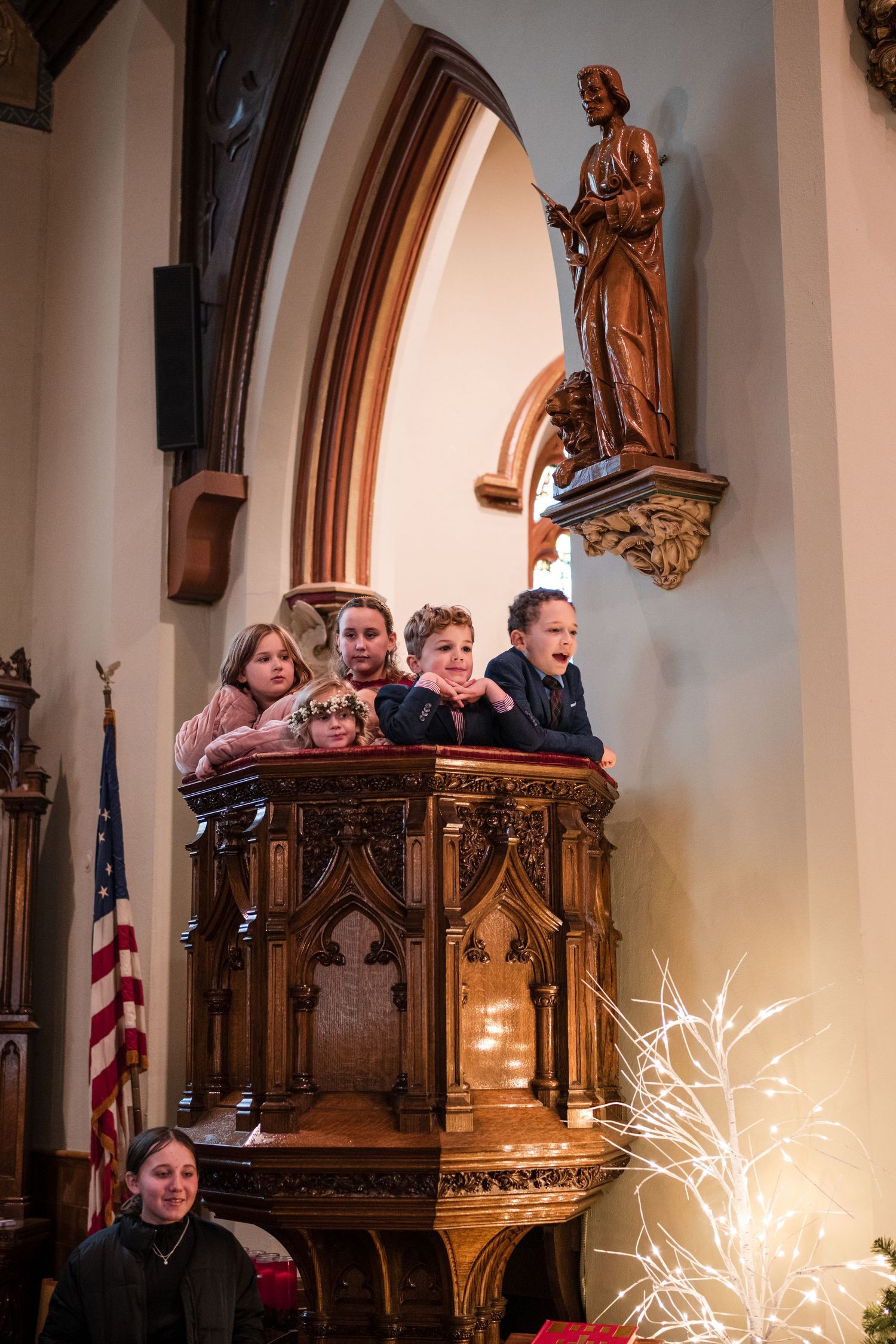 Children and adults gathered in an ornate church pulpit beneath a religious statue.