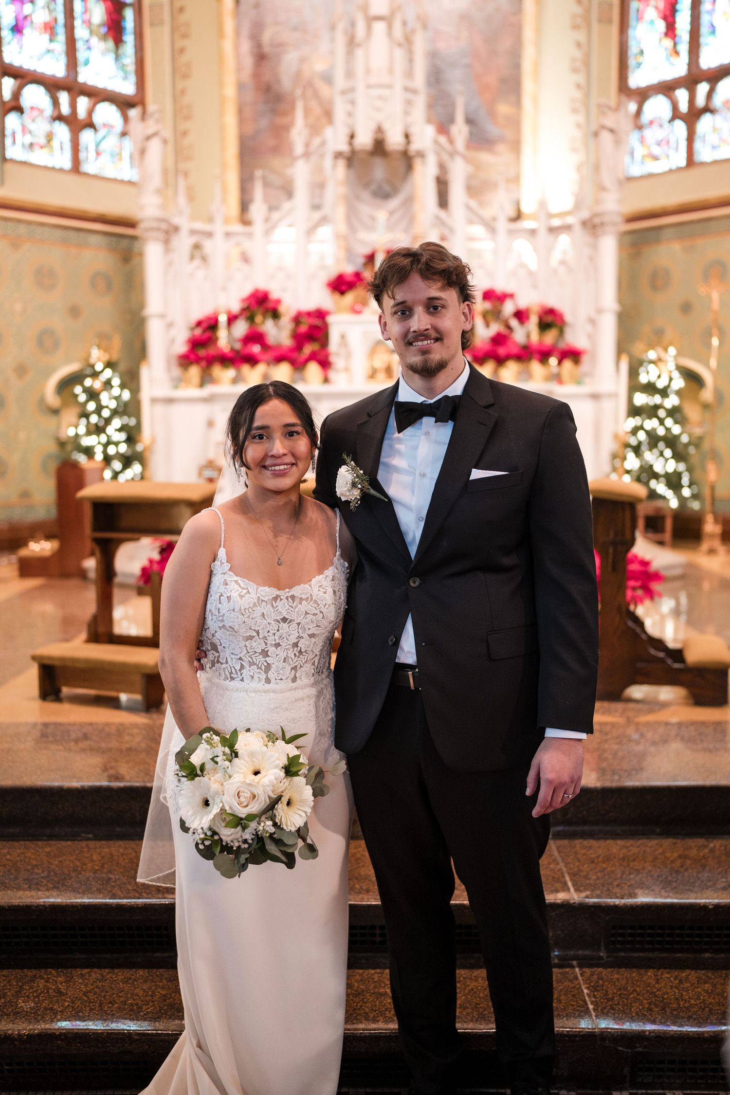 Elegant wedding couple standing at church altar decorated with red poinsettias and twinkling lights.