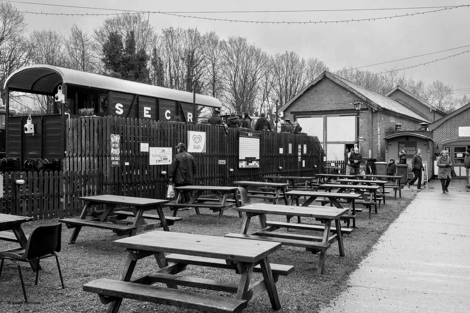 Black and white photo of a vintage train car parked beside outdoor picnic tables at a restaurant or dining venue.