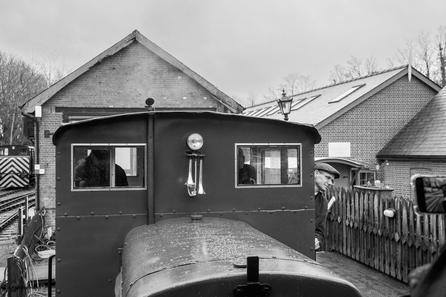 Black and white view from a locomotive cab, showing the exterior of train buildings through the curved windshield glass.