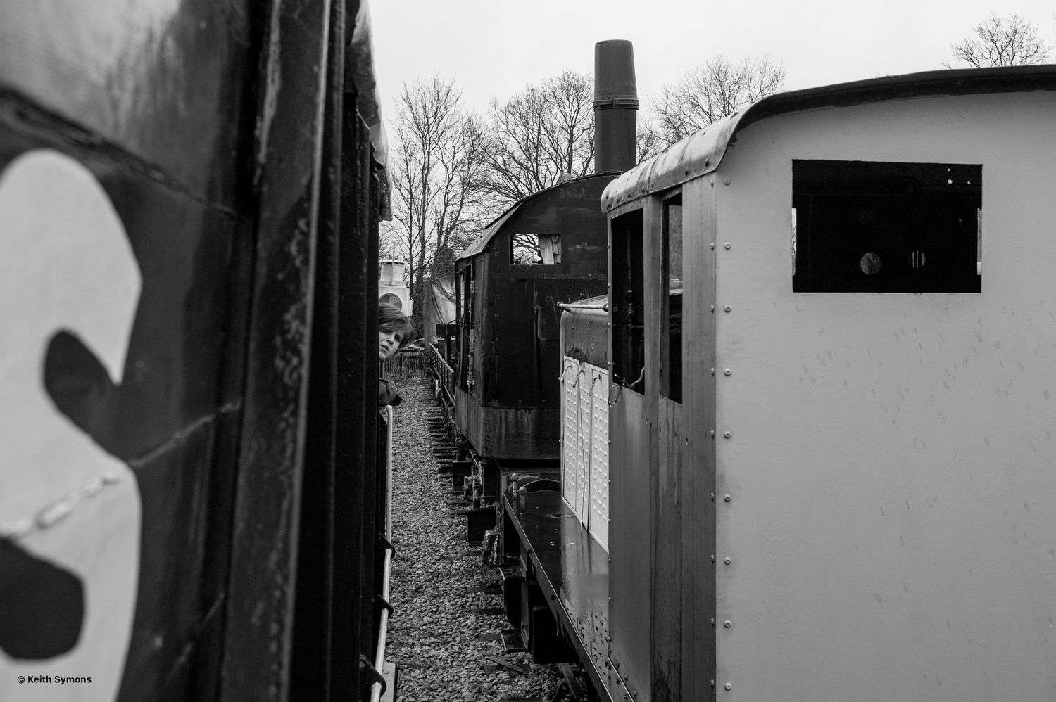 Black and white view of a narrow pathway between vintage railroad cars and equipment at a train yard.