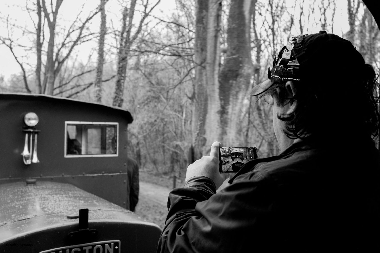 A black and white photo shows someone operating a vintage train engine through a wooded area.