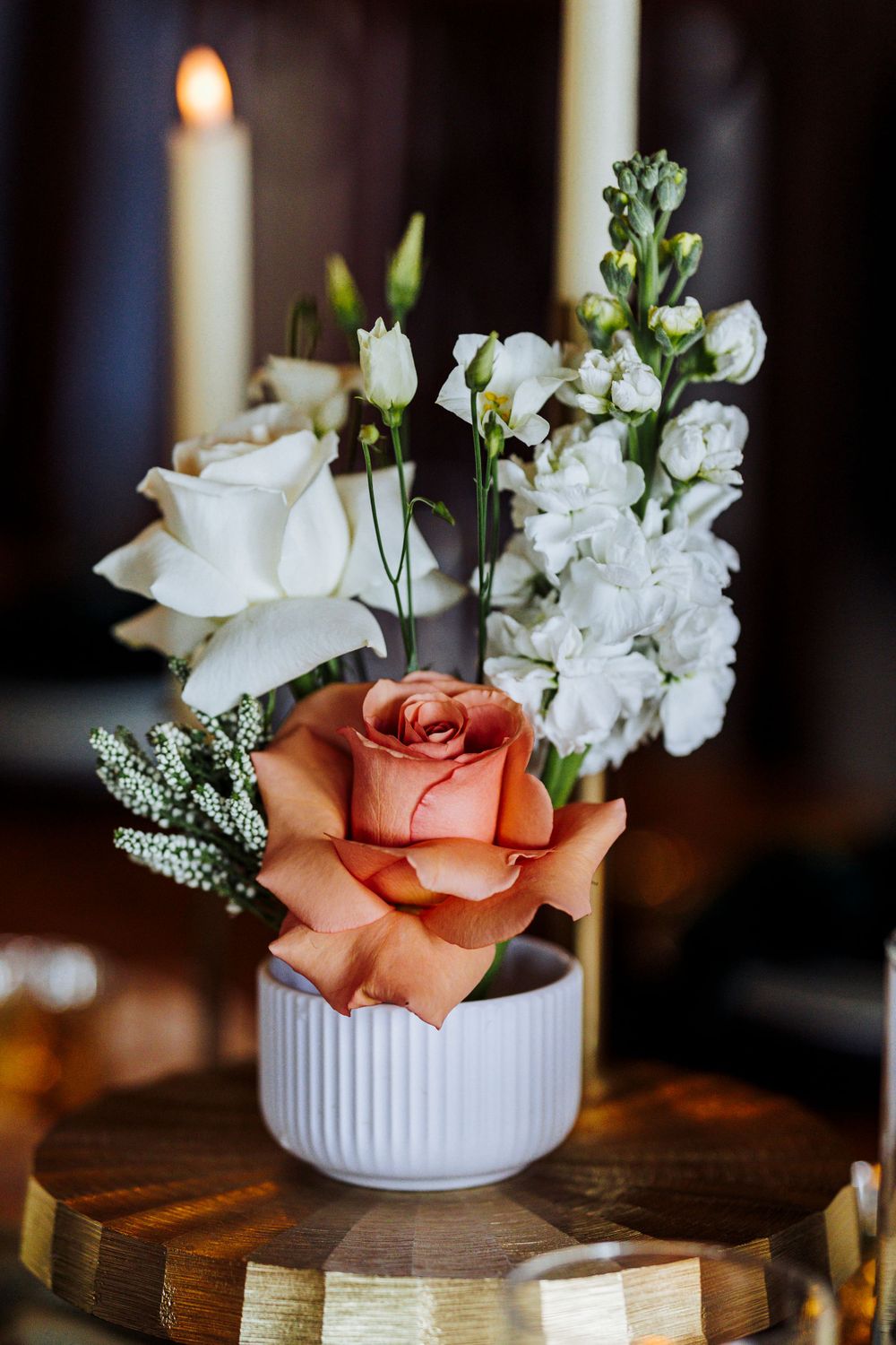 Elegant floral arrangement with a peach rose and white flowers in a ribbed white vase next to a lit candle.