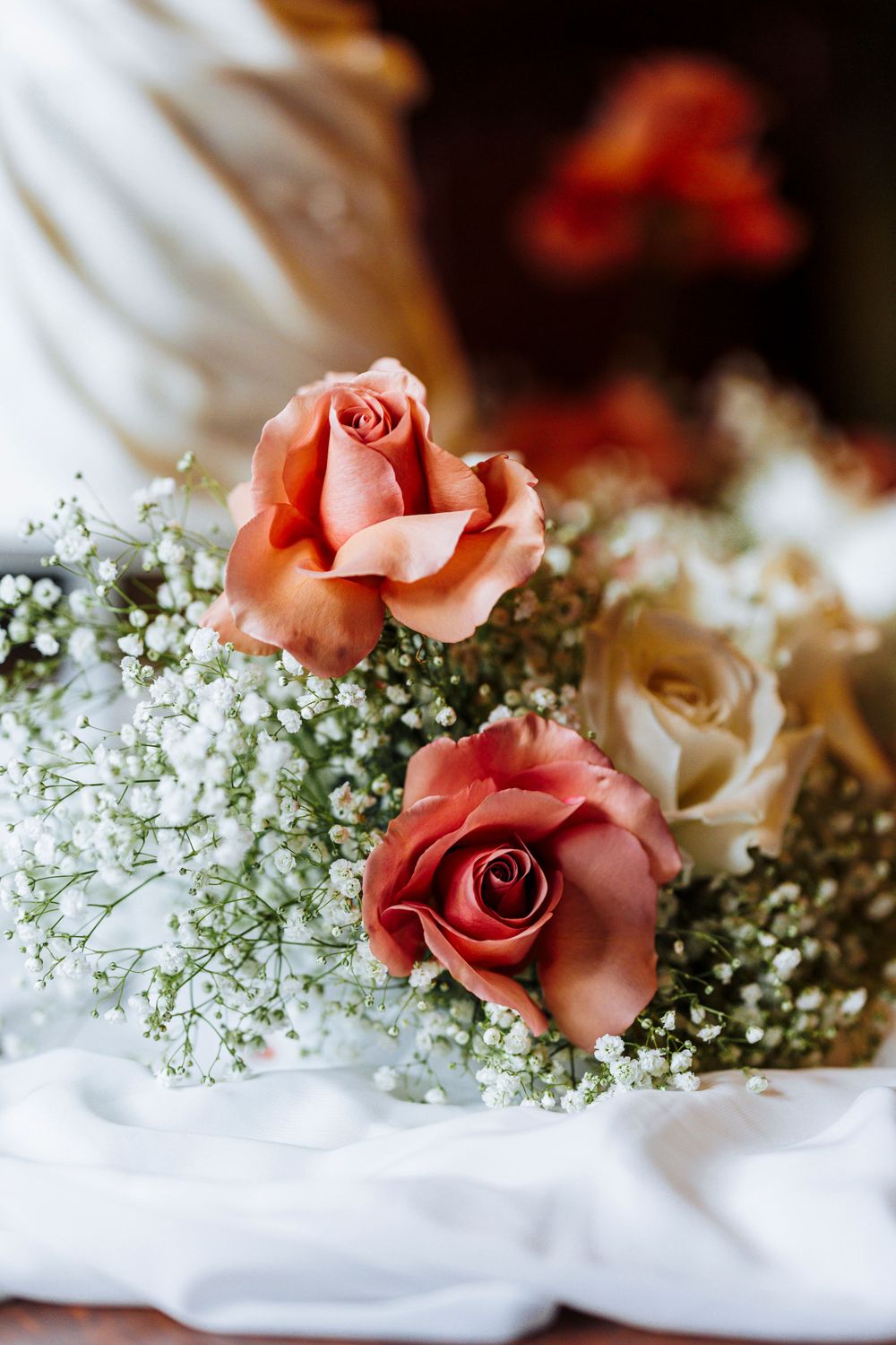 Beautiful coral pink roses nestled among delicate baby's breath flowers against soft white fabric in romantic lighting.