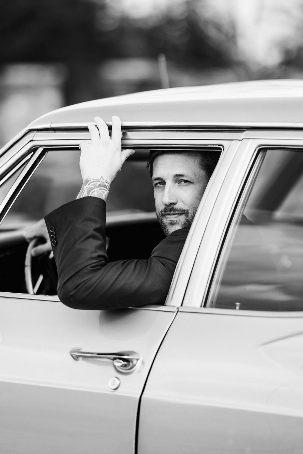A black and white photo of someone looking out the window of a classic car with their arm resting on the door frame.