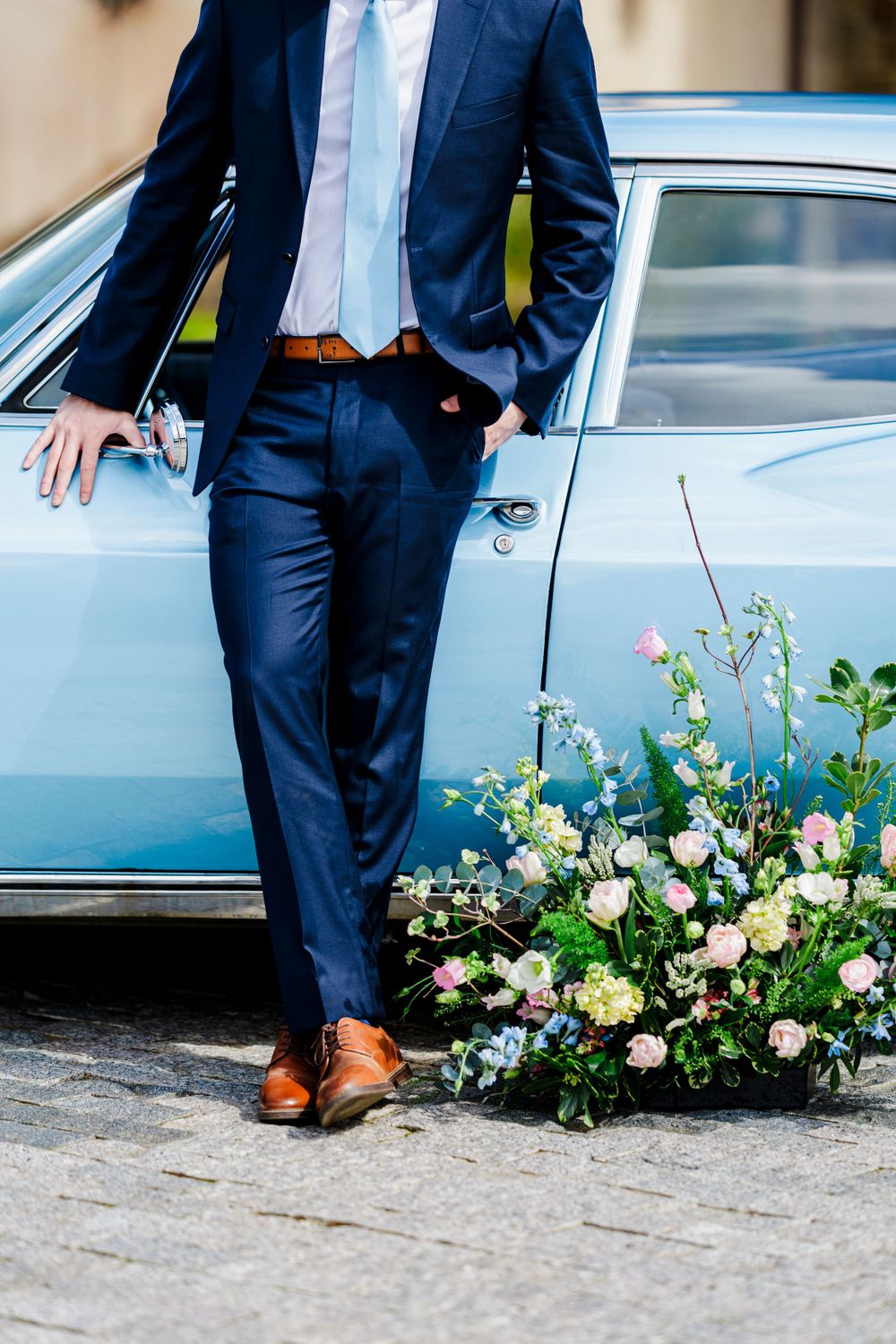 Navy blue suited figure stands beside vintage car with brown leather shoes and floral arrangement at base.