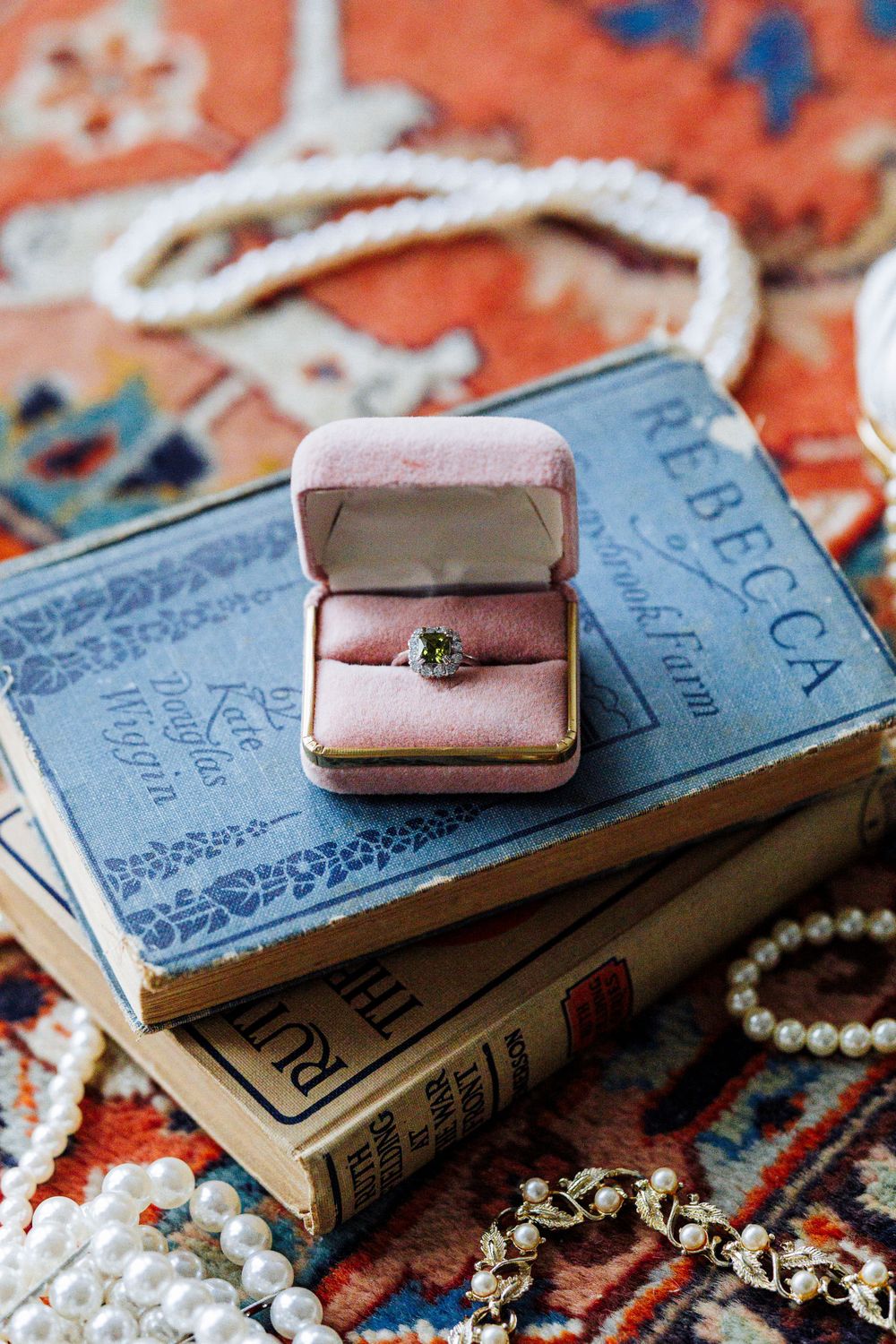 Vintage ring box with engagement ring displayed on antique books and pearls against an orange patterned rug.