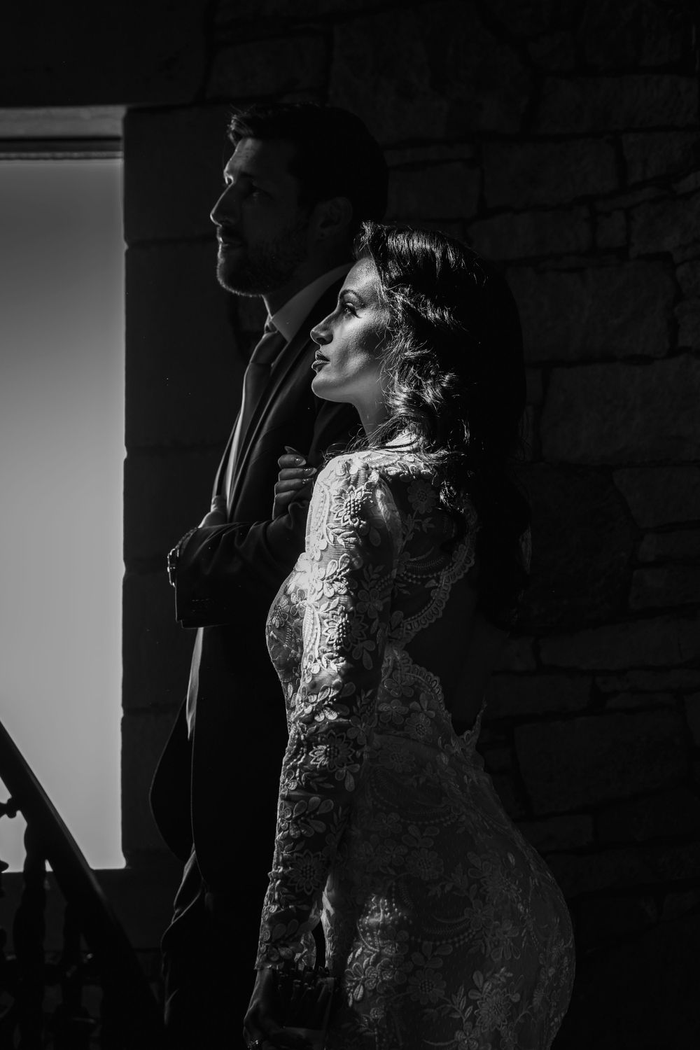 A dramatic black and white portrait of a couple in formal attire standing against a brick wall in low-key lighting.