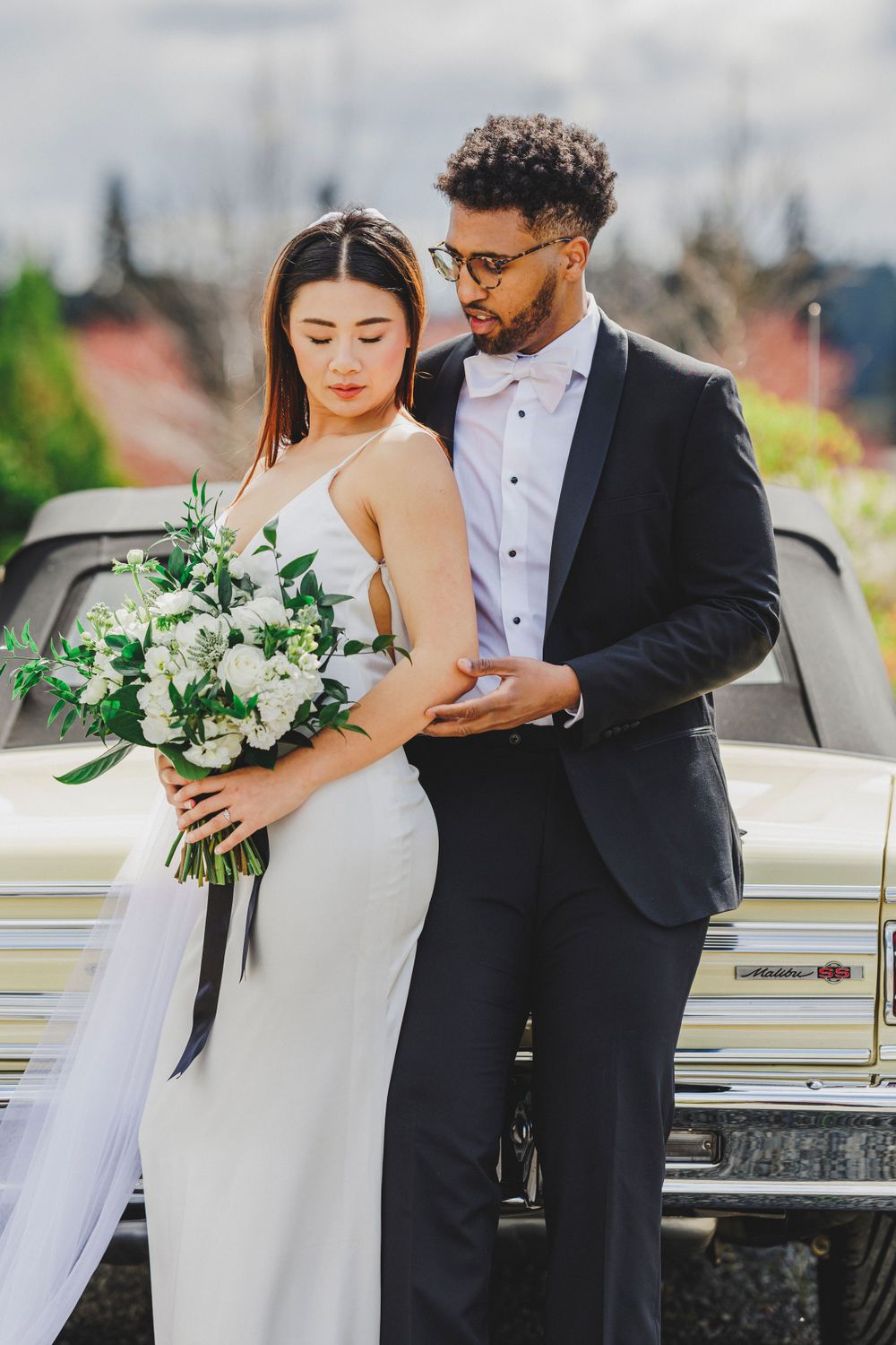 Elegant wedding couple poses with white floral bouquet next to vintage car in formal attire.