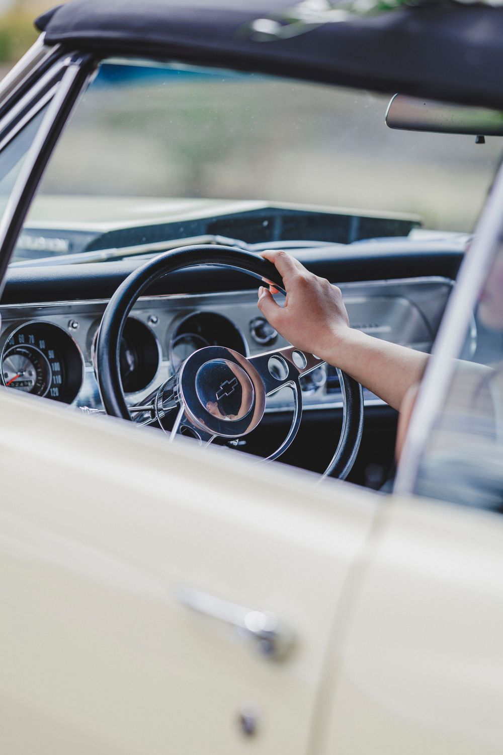 View through car window showing hand on vintage steering wheel of classic car with retro dashboard gauges.