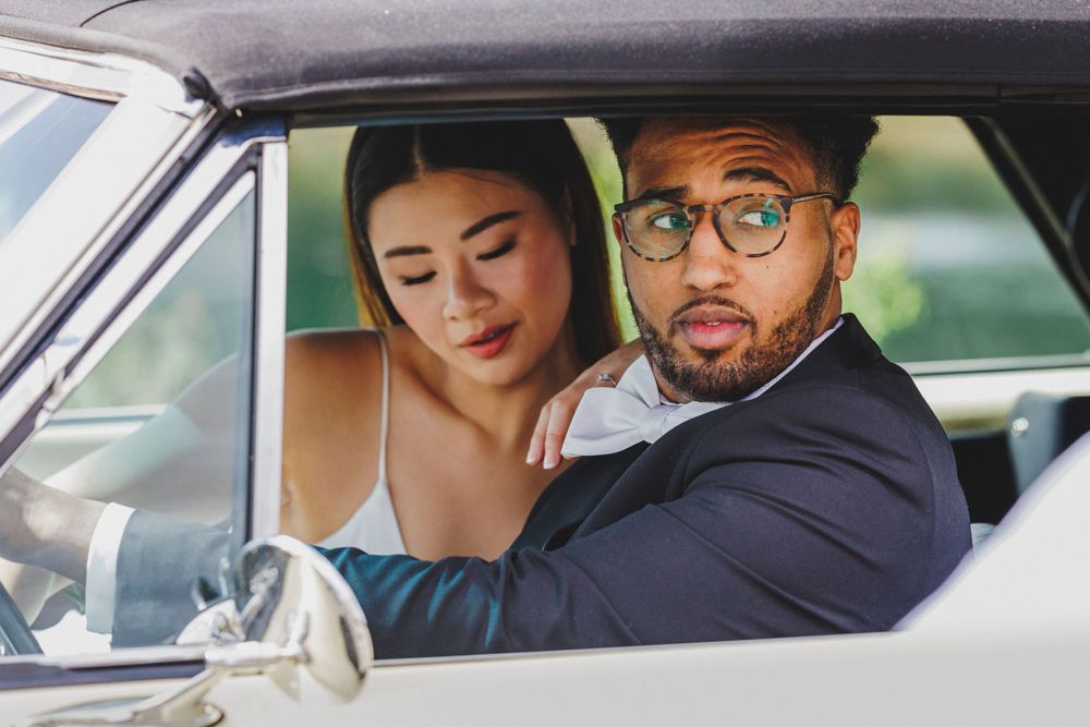 A couple dressed in formal attire sit together in a vintage car, with the driver playfully making a surprised expression.