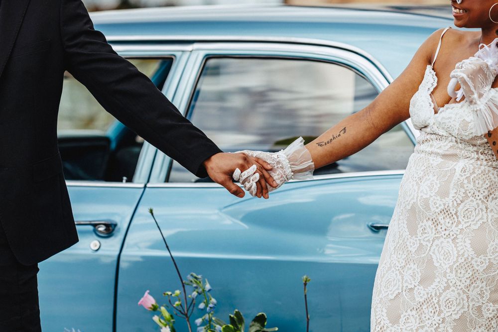 Two hands clasped together against backdrop of vintage blue car at wedding celebration.