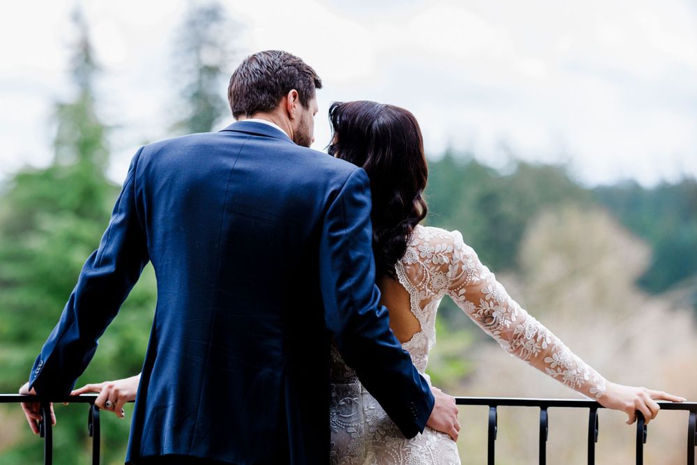 A newlywed couple in formal attire embrace while overlooking a scenic mountain view from a balcony railing.