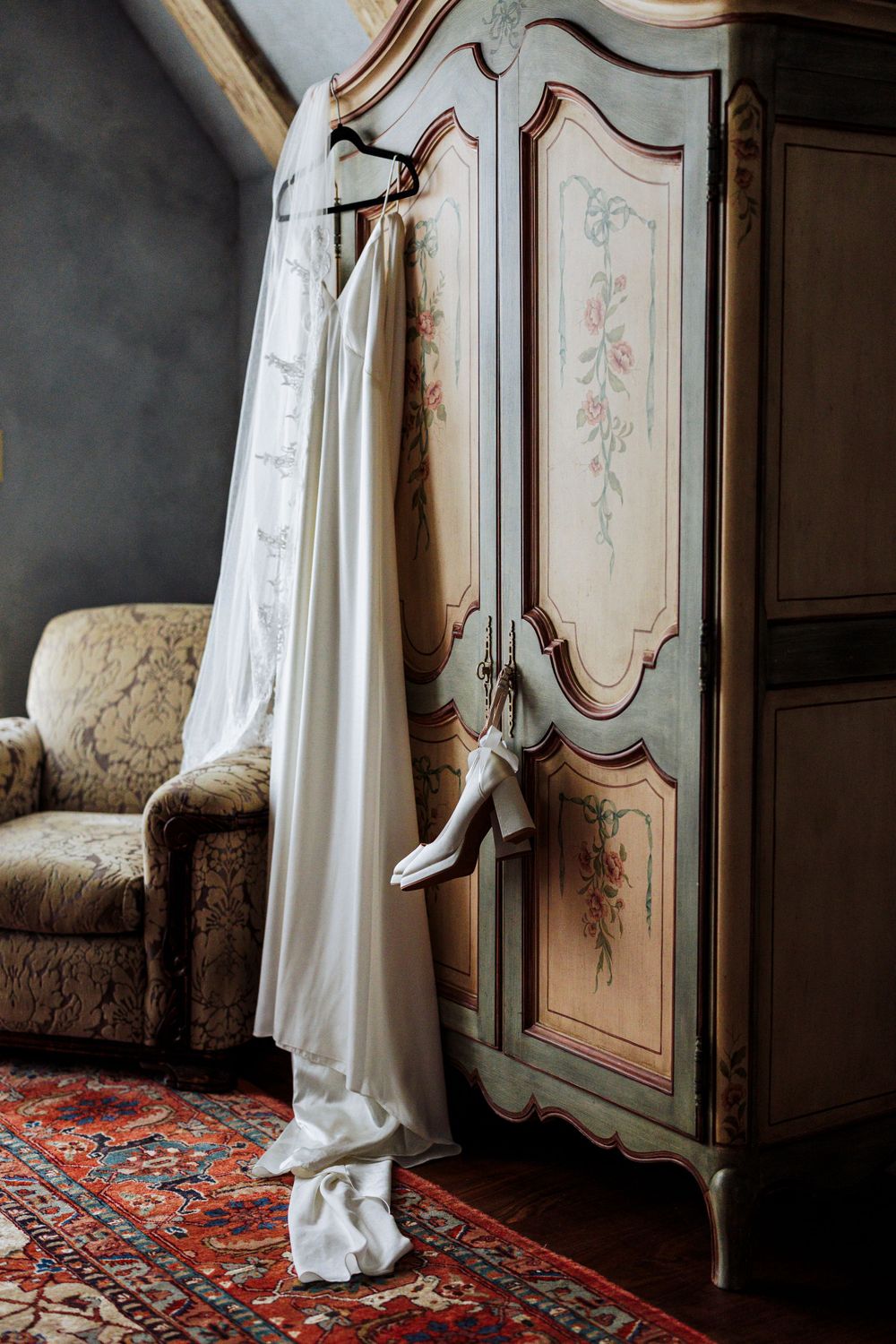 A white wedding dress hangs from an elegant vintage armoire with floral designs, beside a plush chair on a Persian rug.