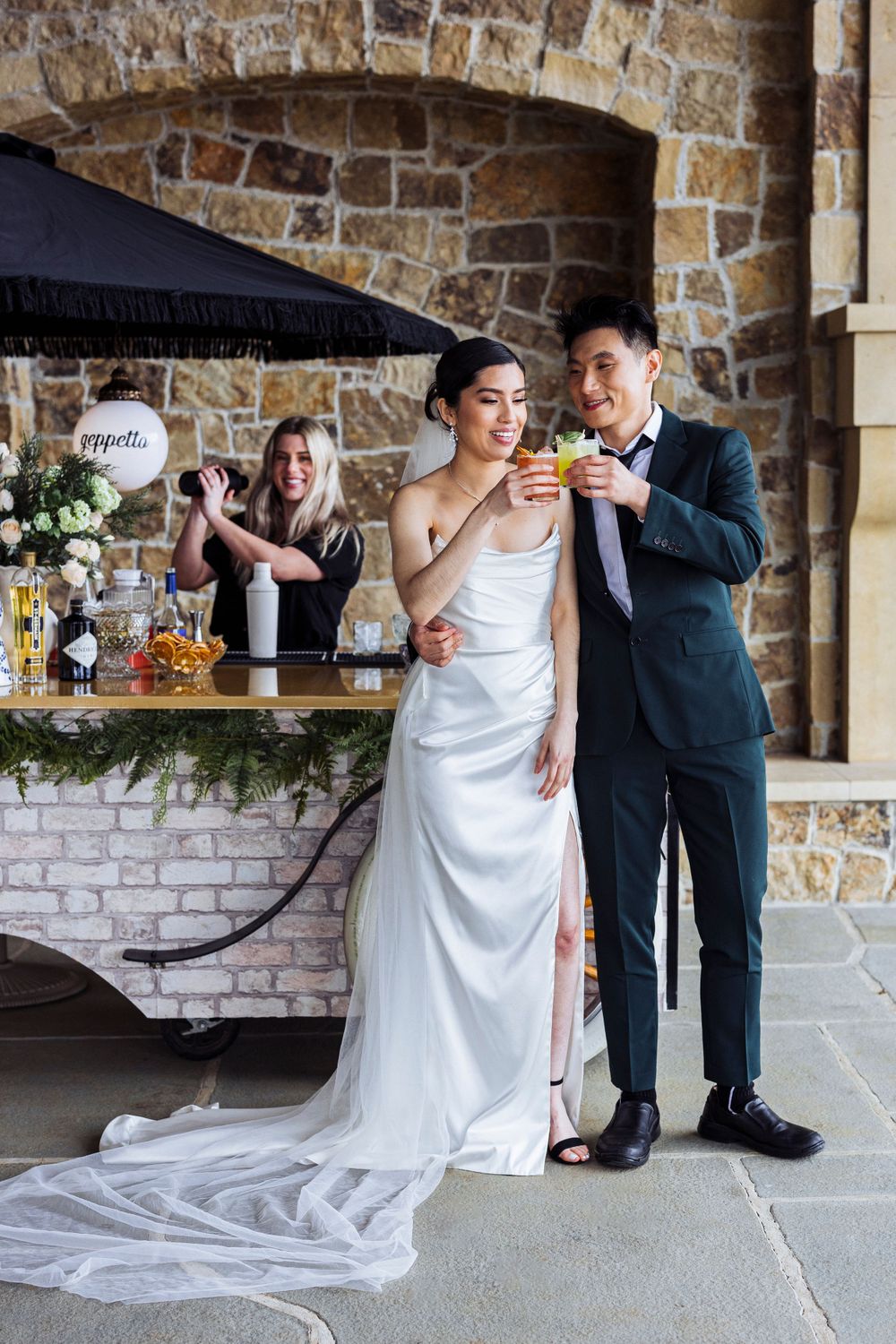 A bride in a white gown and groom in dark suit share a drink at a rustic stone venue with bar in background.
