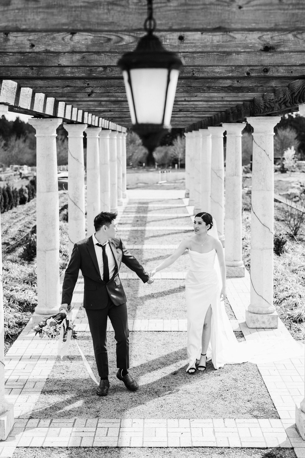 Black and white photo of couple walking under elegant columned walkway with hanging lantern at a garden venue.