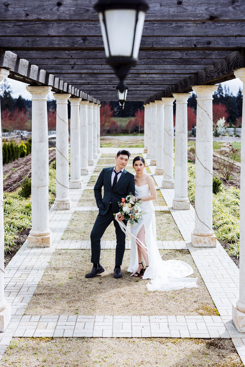 A couple poses under a covered walkway with white columns and overhead lanterns in an elegant outdoor setting.