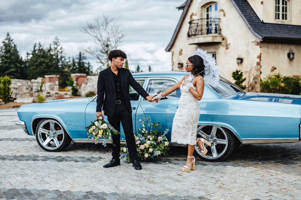 A couple poses with a classic bright blue vintage car outside an elegant estate on a cloudy day.