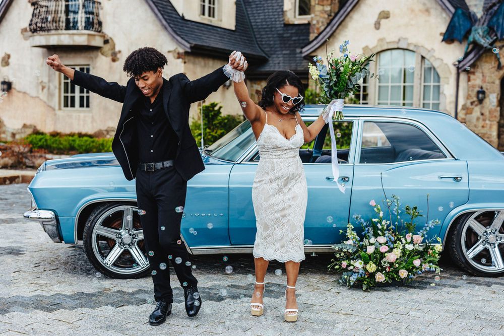 Two people jump joyfully in front of a vintage light blue car decorated with flowers in an outdoor setting.