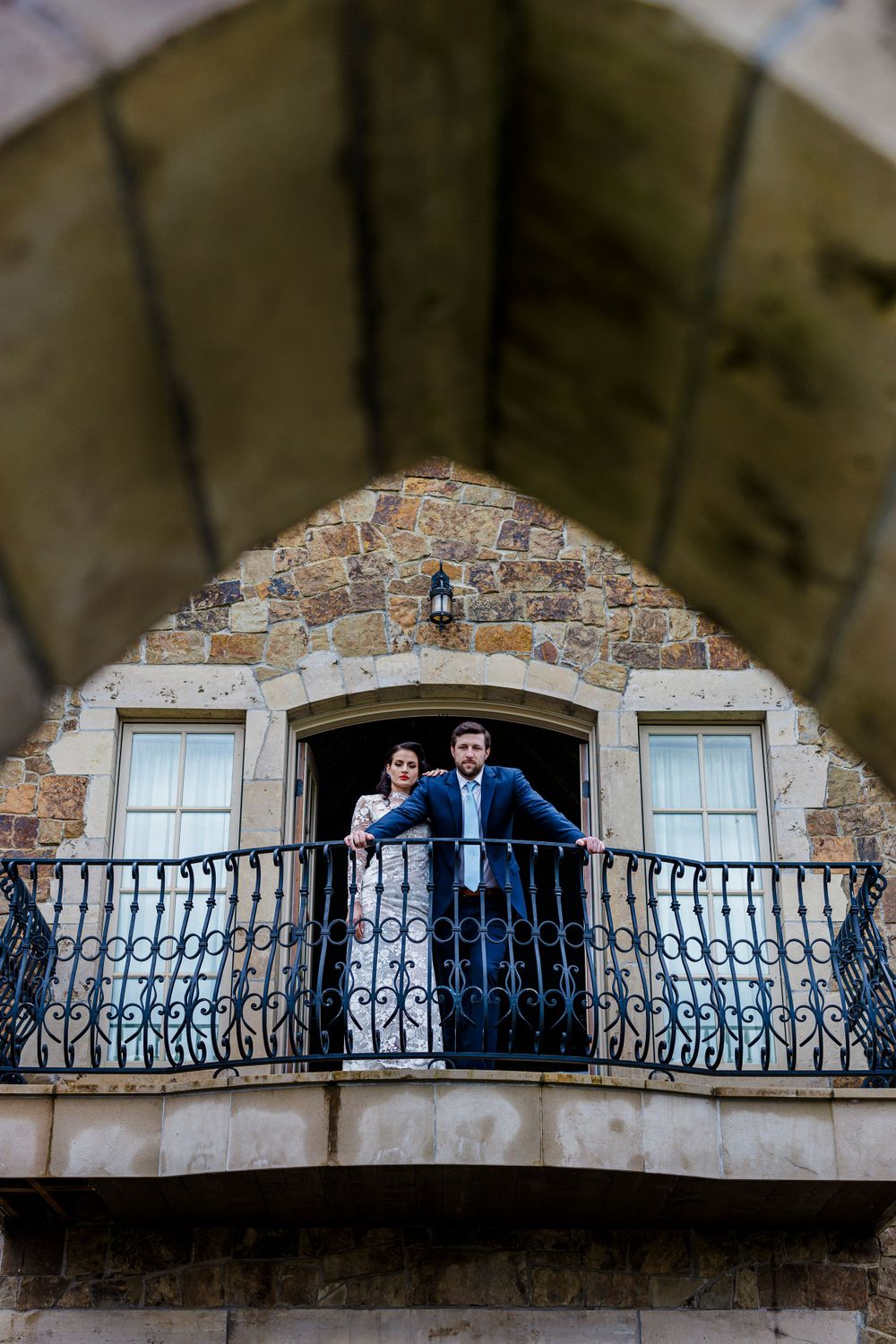 A couple stands together on an ornate stone balcony with wrought iron railings of a rustic building.