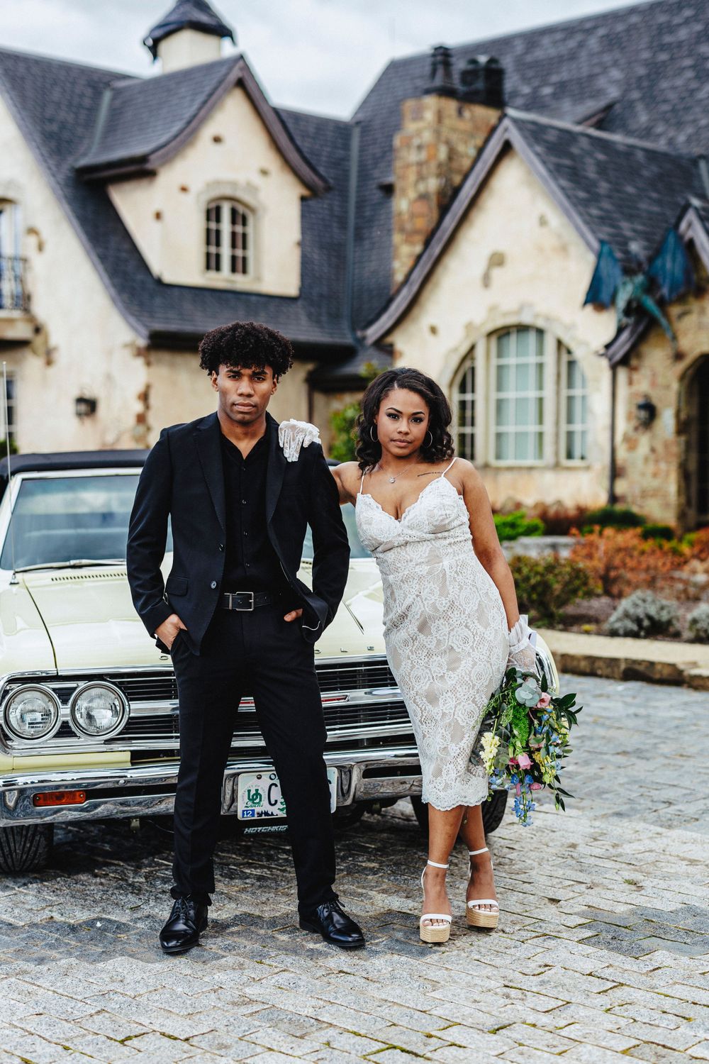 A couple in wedding attire poses in front of a vintage car and French country-style house with stone exterior.