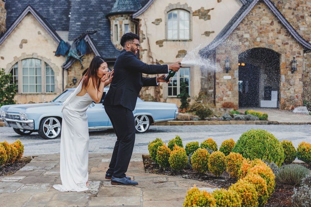 A couple strikes energetic poses in front of a stone cottage with vintage car and manicured landscaping in background.