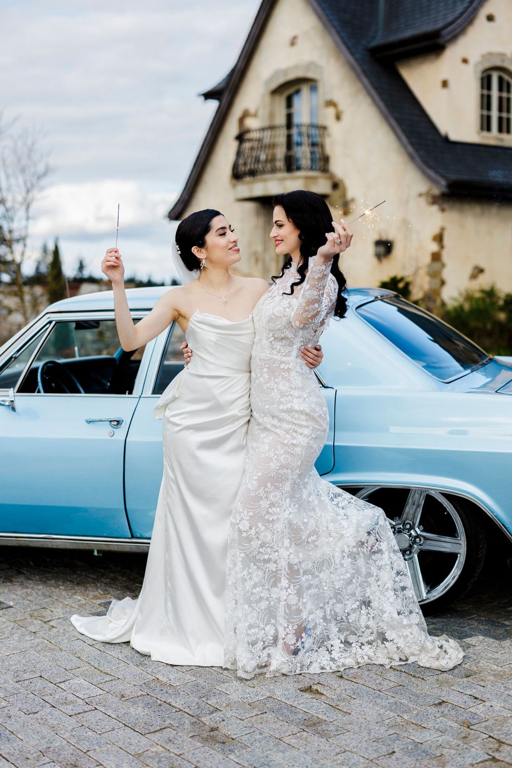 Two brides in white wedding dresses share a joyful moment together beside a classic light blue car.
