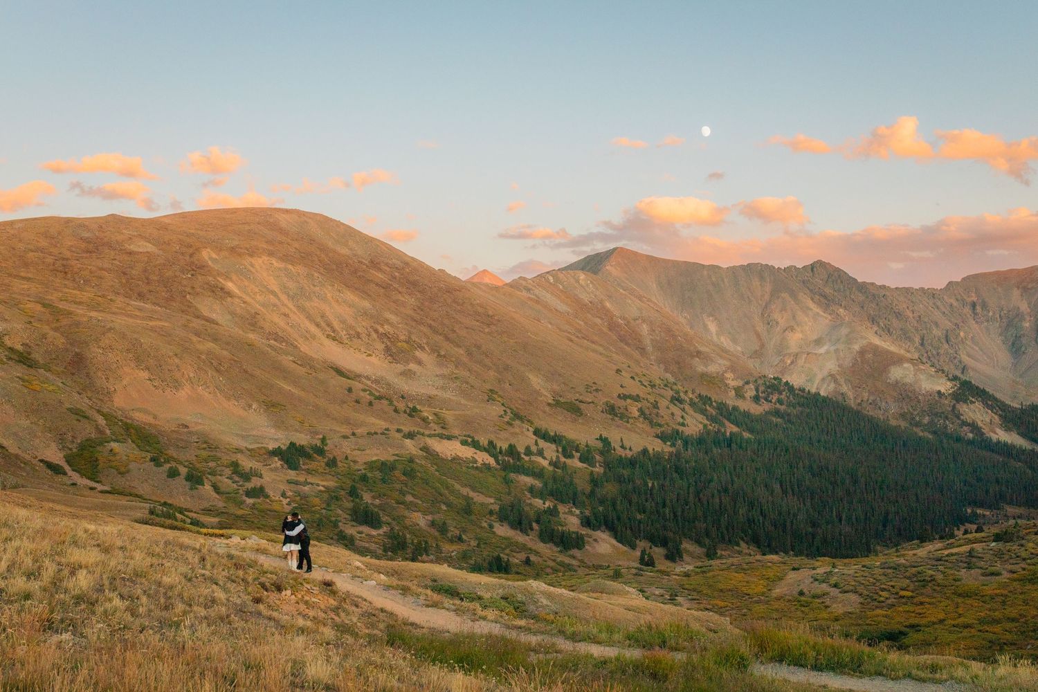 A hiker explores a winding trail through golden mountains at sunset, with forest-covered slopes below and pink clouds above.