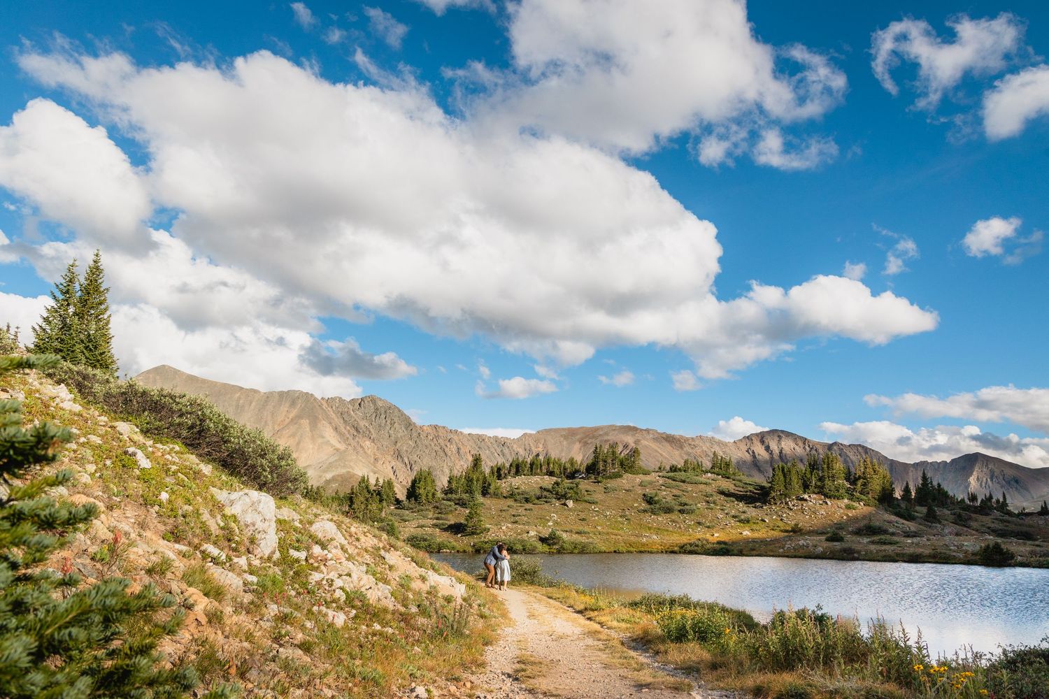 Hiking trail winds along an alpine lake with rugged mountain peaks and fluffy white clouds in the blue sky.