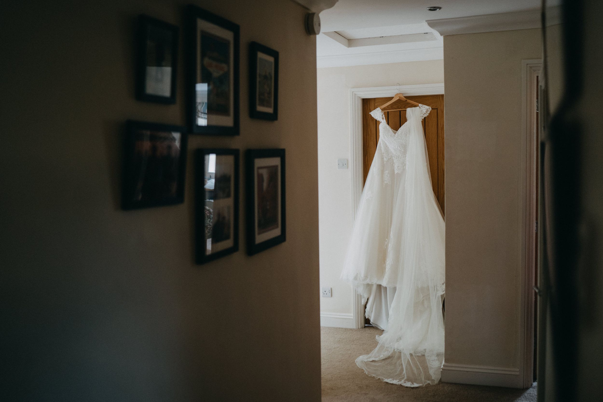 A white wedding dress hangs elegantly in a doorway with framed photos on the wall in the foreground.