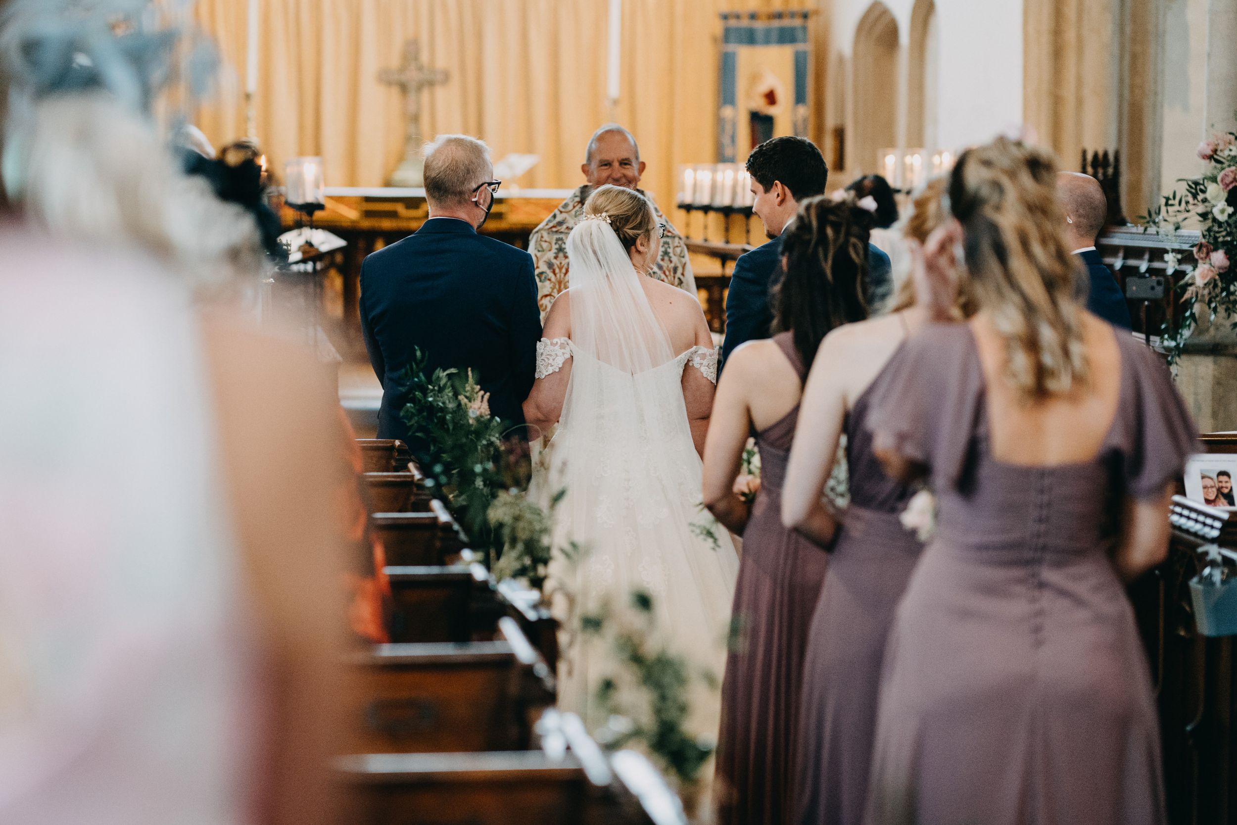 Wedding ceremony taking place inside a traditional church with purple-clad bridesmaids lining the aisle.