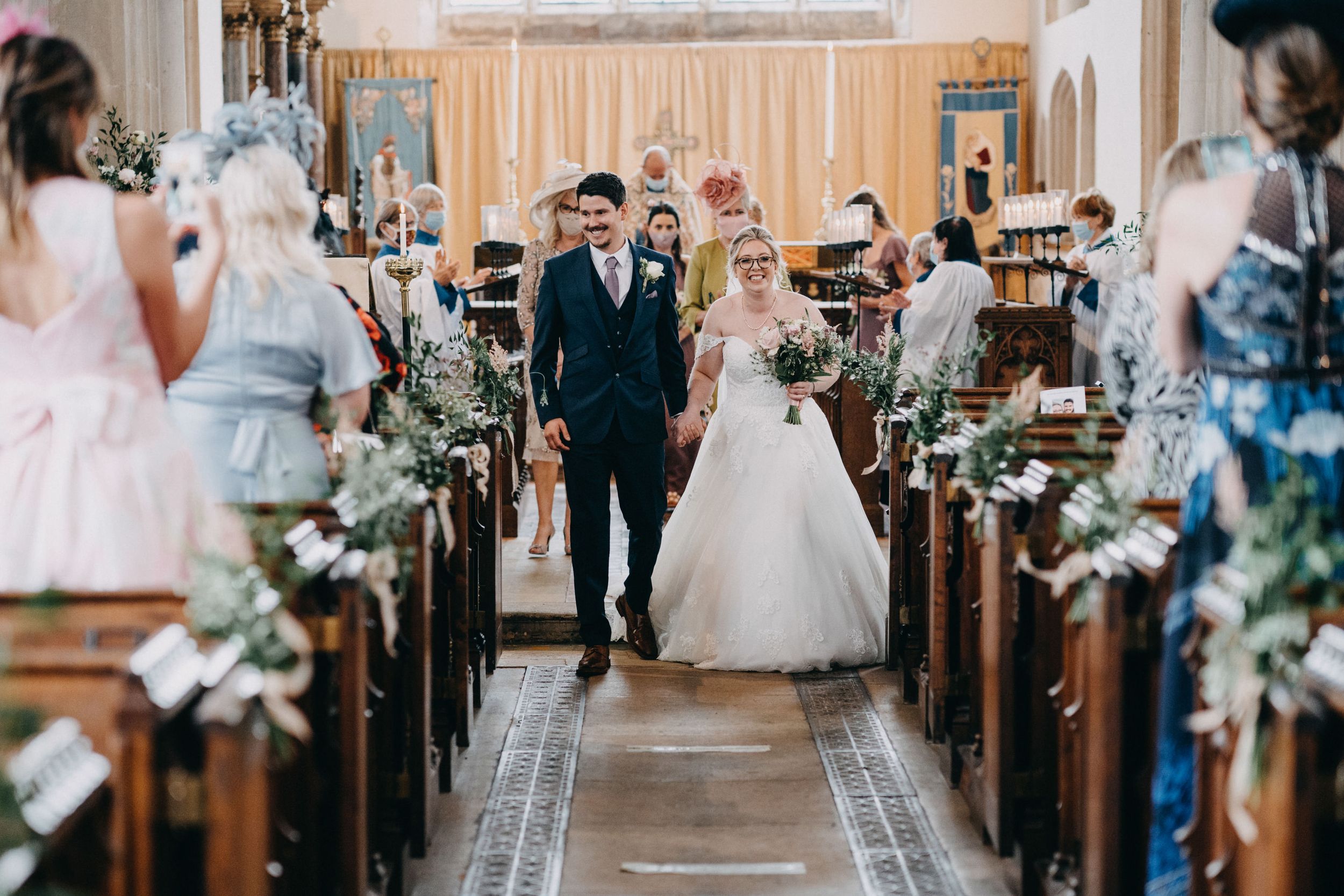Beautiful church wedding ceremony with couple walking down flower-decorated aisle while guests watch from wooden pews.