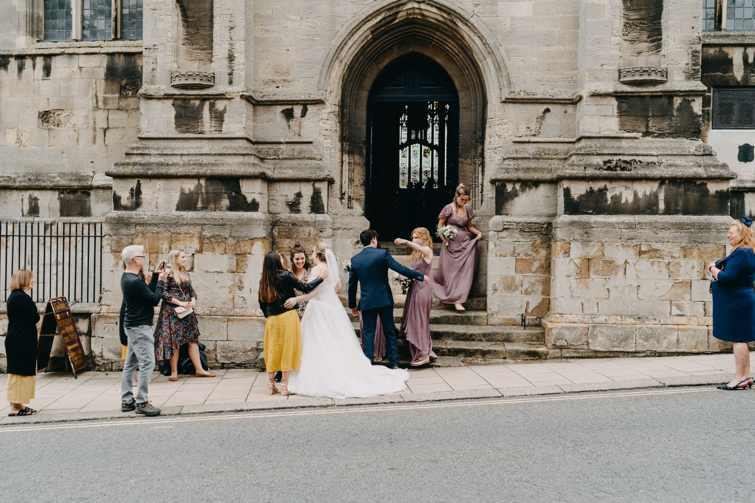 A wedding party gathers at the stone entrance of a historic building as onlookers watch nearby.