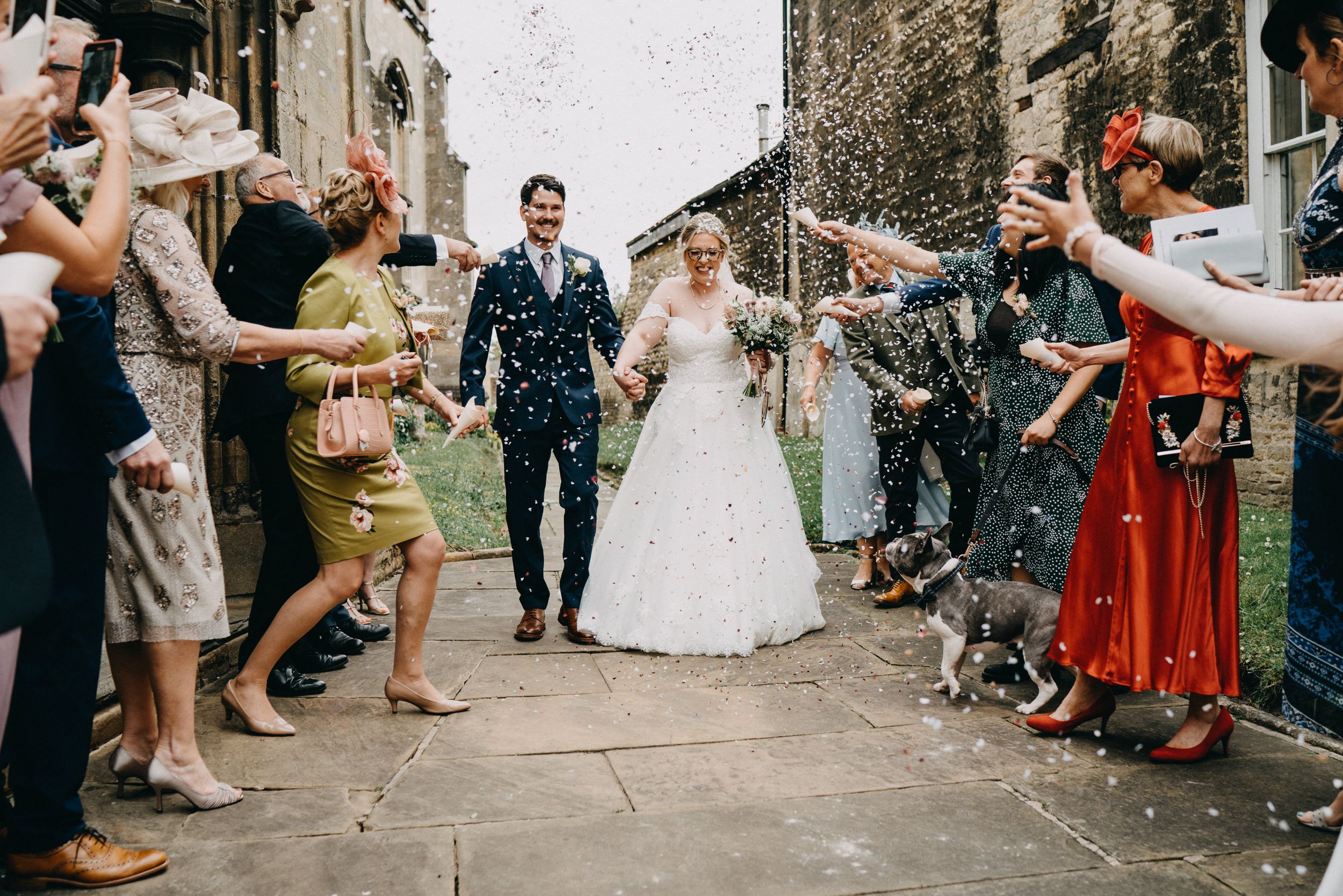 Wedding guests throw confetti over newlyweds during a joyful celebration outside a stone church building.