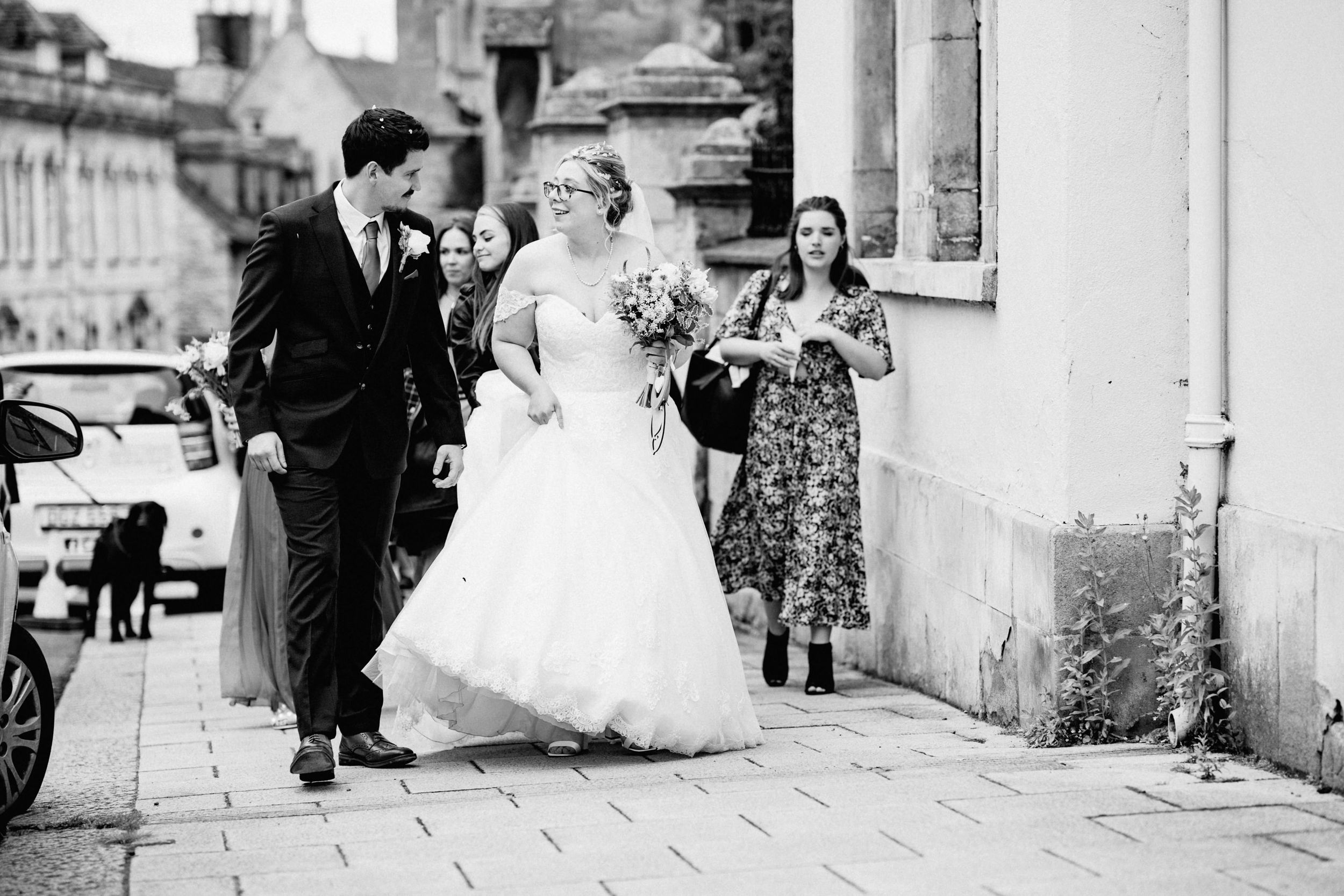 Black and white wedding photo showing a couple in formal attire walking along a sidewalk with a third person watching.