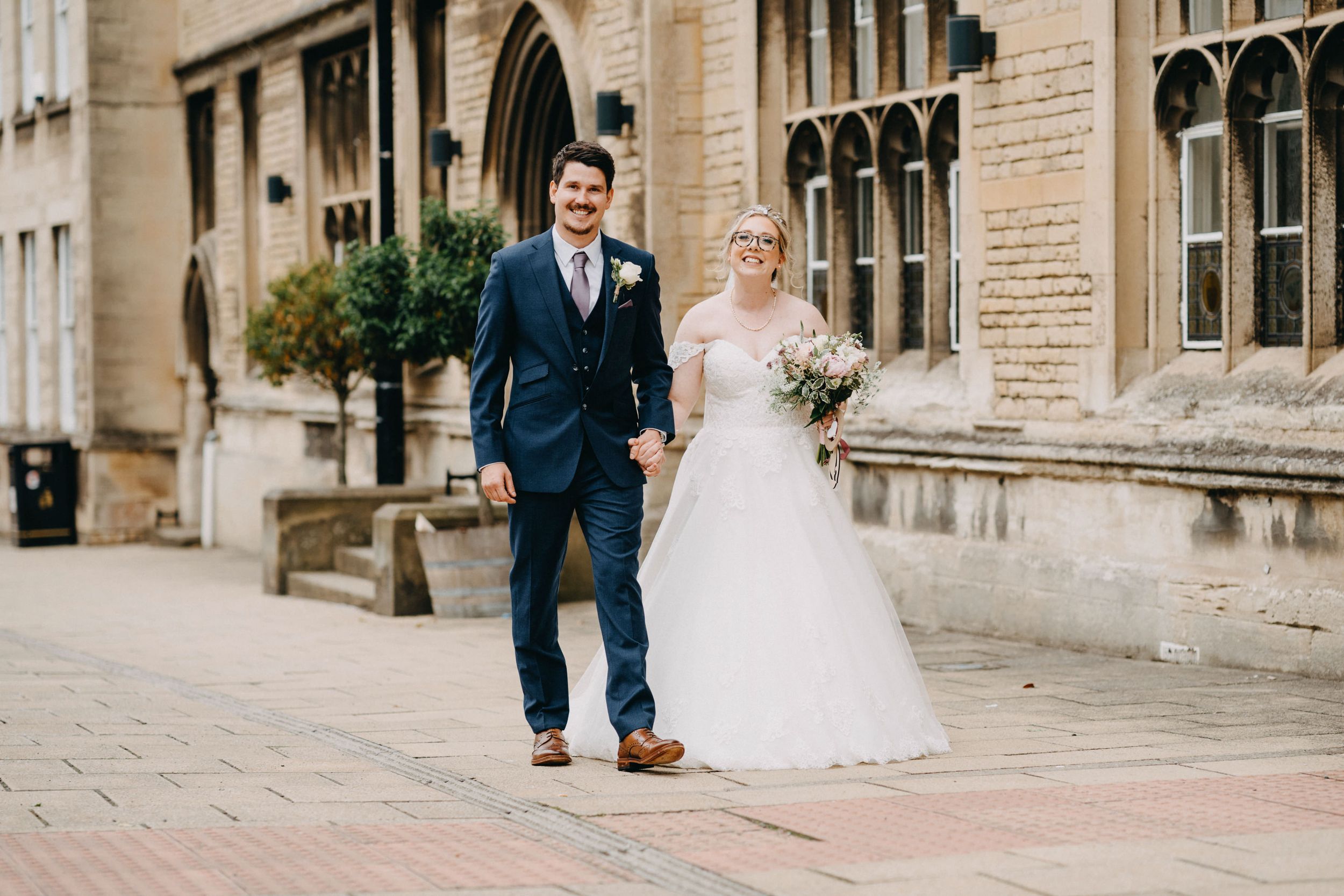 A couple walks hand in hand outside a historic stone building on their wedding day, with bride in white gown and groom in navy suit.
