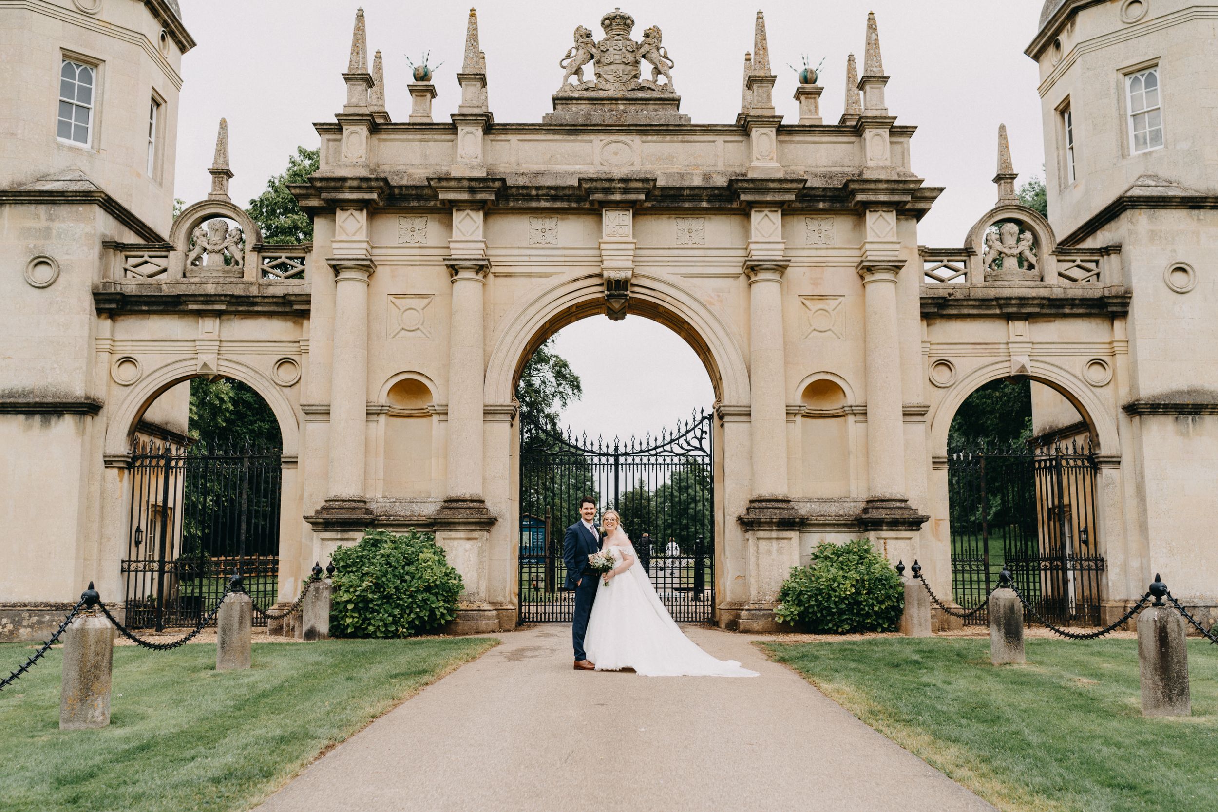 A couple stands in the ornate archway entrance of a grand historic estate with stone spires and decorative architecture.