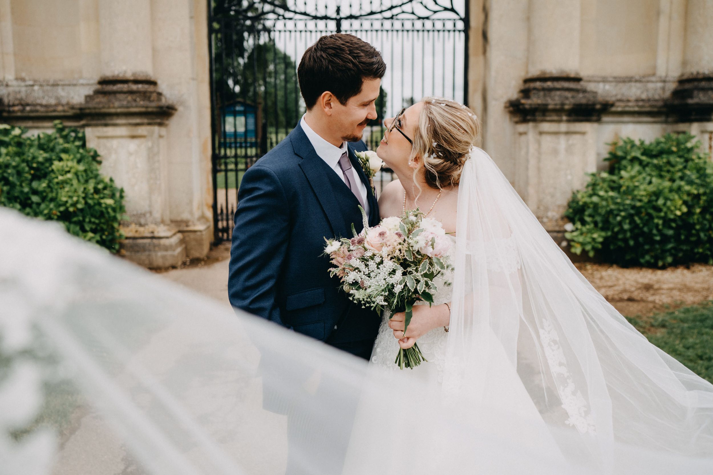 A bride and groom share a romantic moment in front of an ornate iron gate while her veil flows in the breeze.