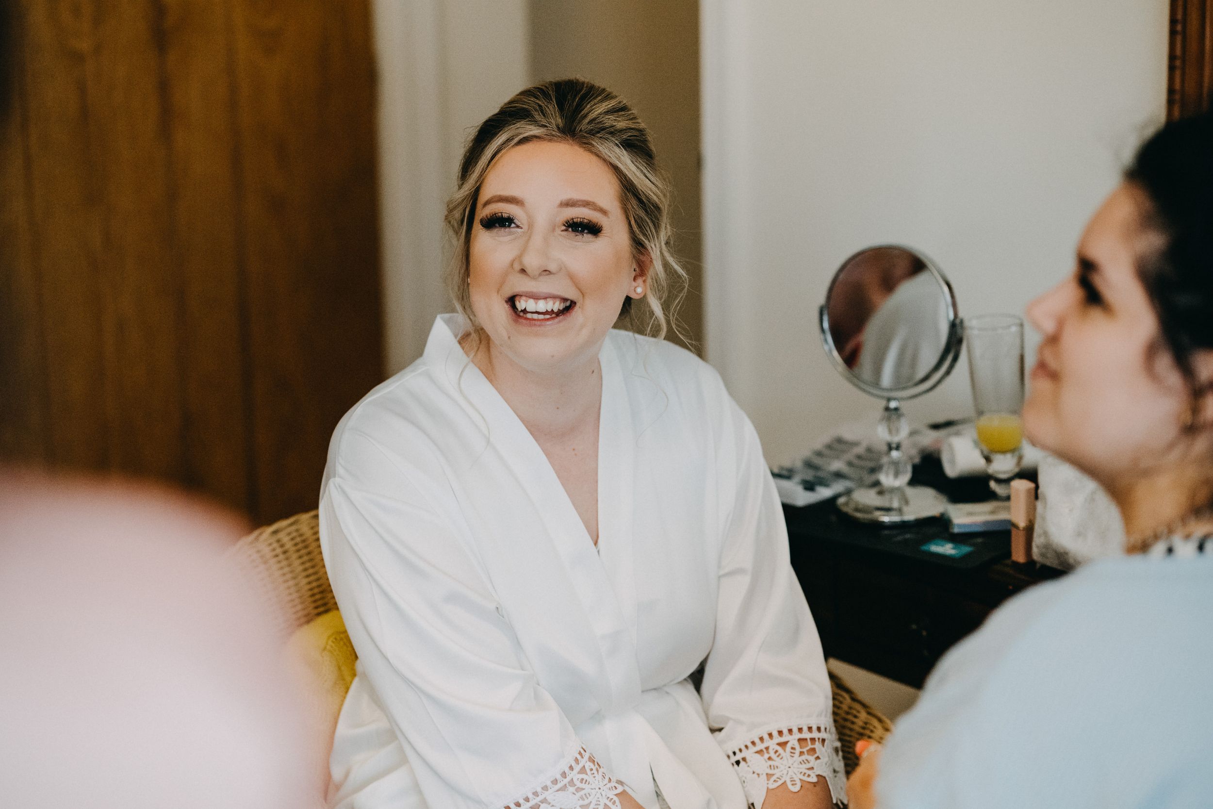 A person wearing a white robe with lace trim smiles while getting ready in a dressing room.