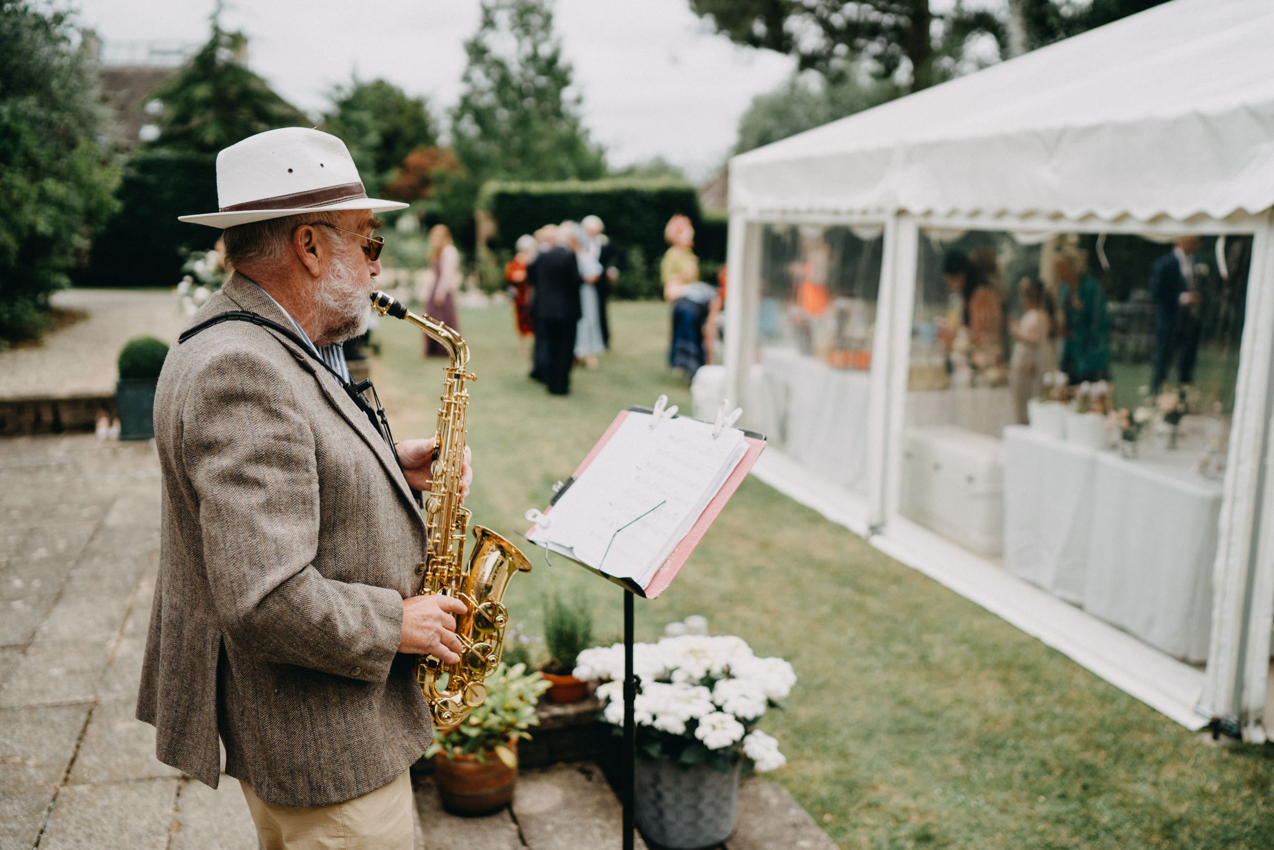 A saxophone player in a tweed jacket performs next to a white wedding tent on a lawn.