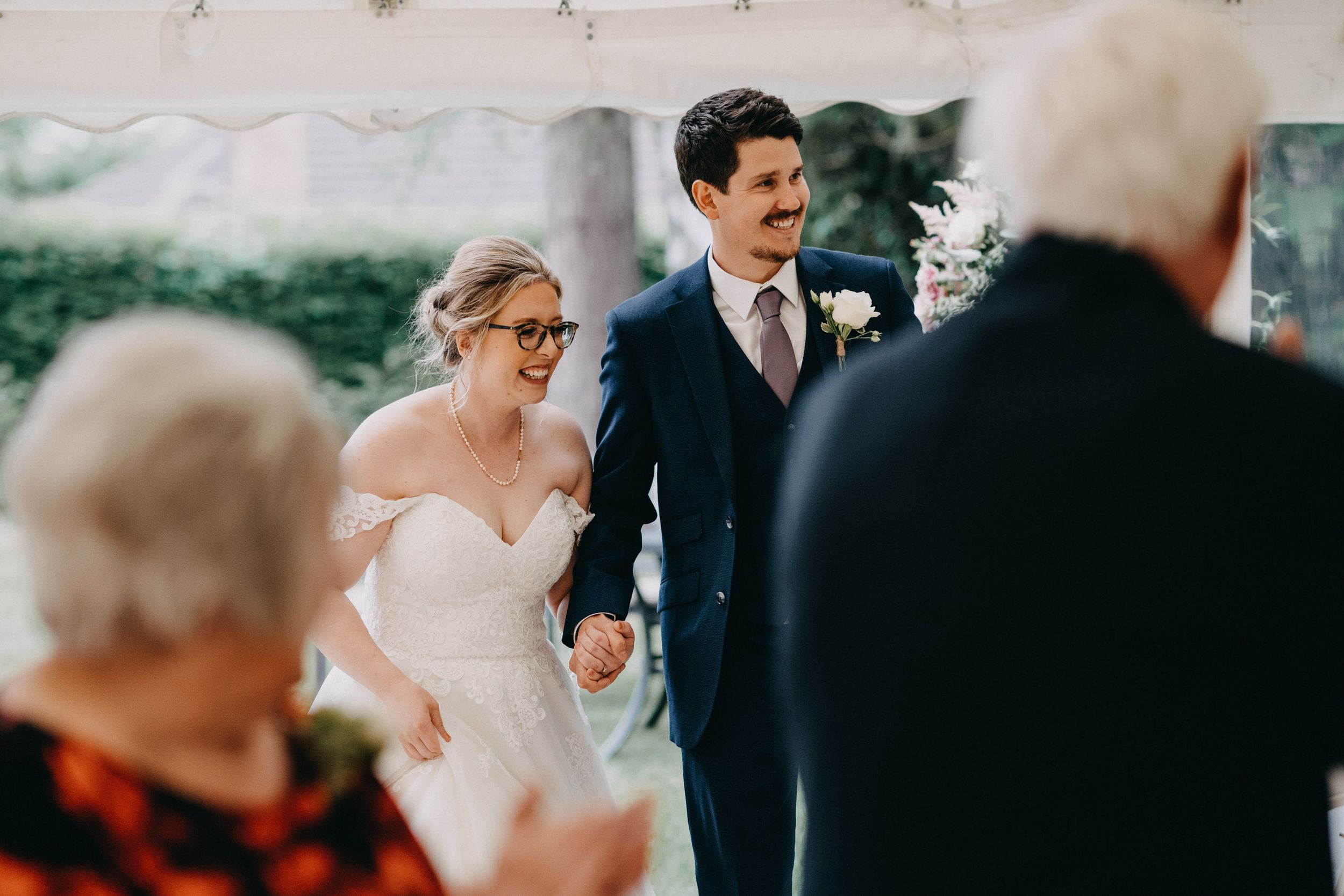 Newlyweds hold hands during outdoor wedding ceremony while guests look on from foreground.