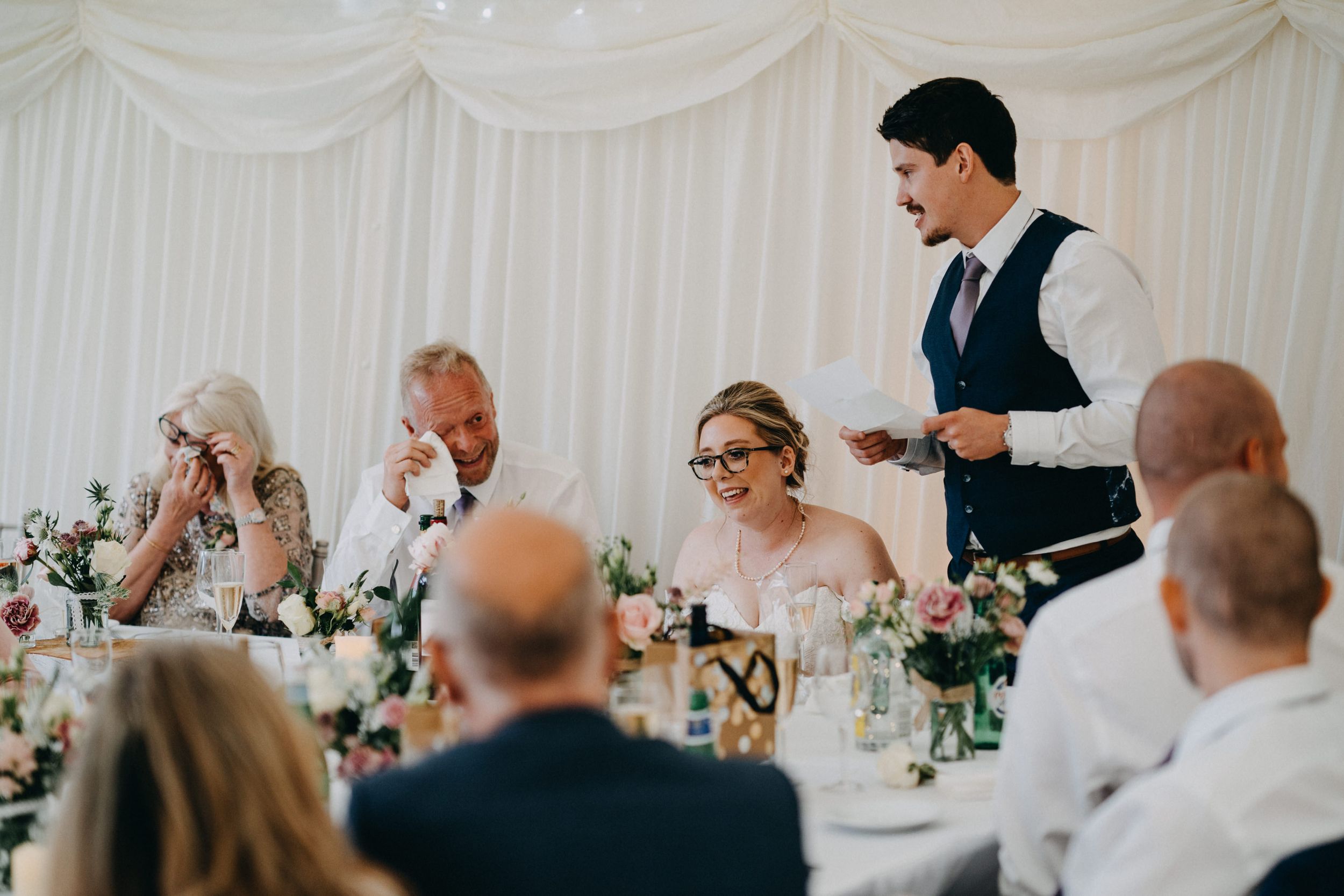 Wedding reception guests seated at decorated table in white marquee while server stands to take orders.