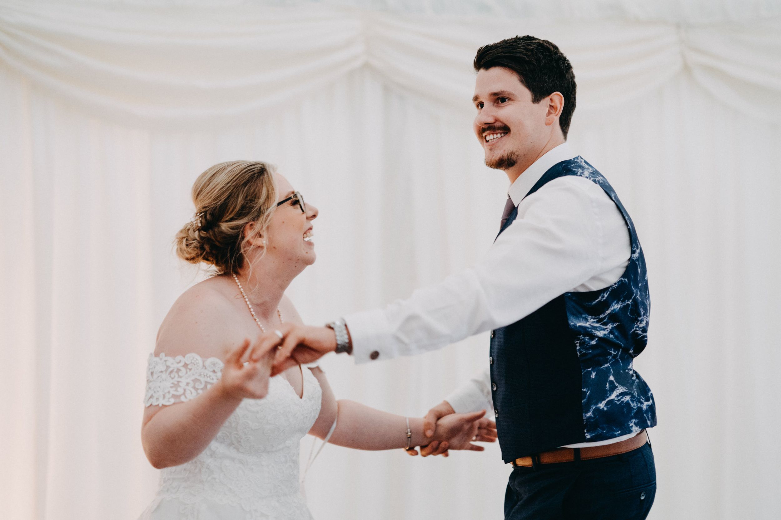 A couple dances joyfully together during a wedding reception in a white-draped venue.