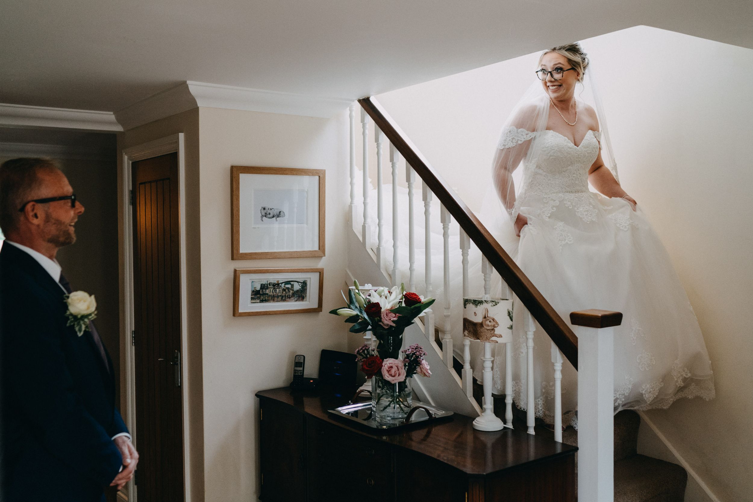 A person in a flowing white wedding dress descends a staircase while someone waits near a floral arrangement below.